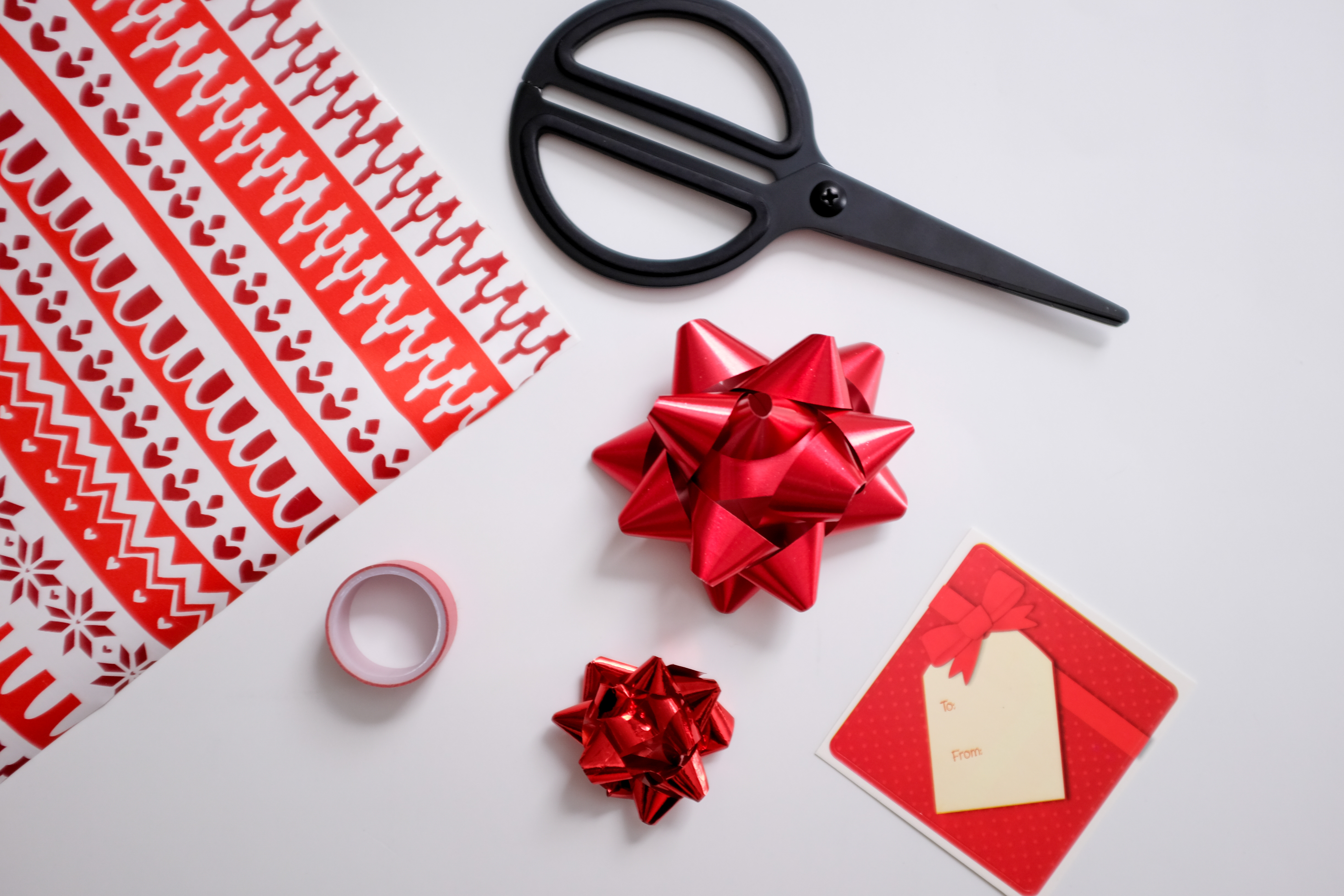 A flatlay of holiday gift wrap materials: gift wrap, scissors, bows, tape, and a to/from tag