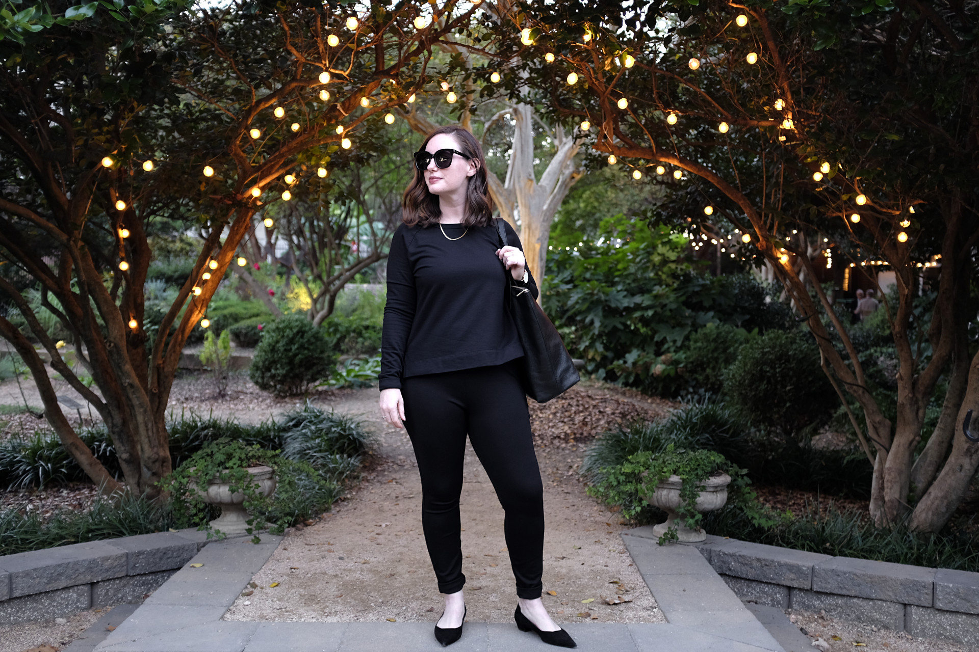 A white woman stands in a garden strung with fairy lights. She is wearing black from head to toe: a black sweatshirt, black pants, black shoes, black sunglasses, and is carrying a black bag. She is looking off to her right.