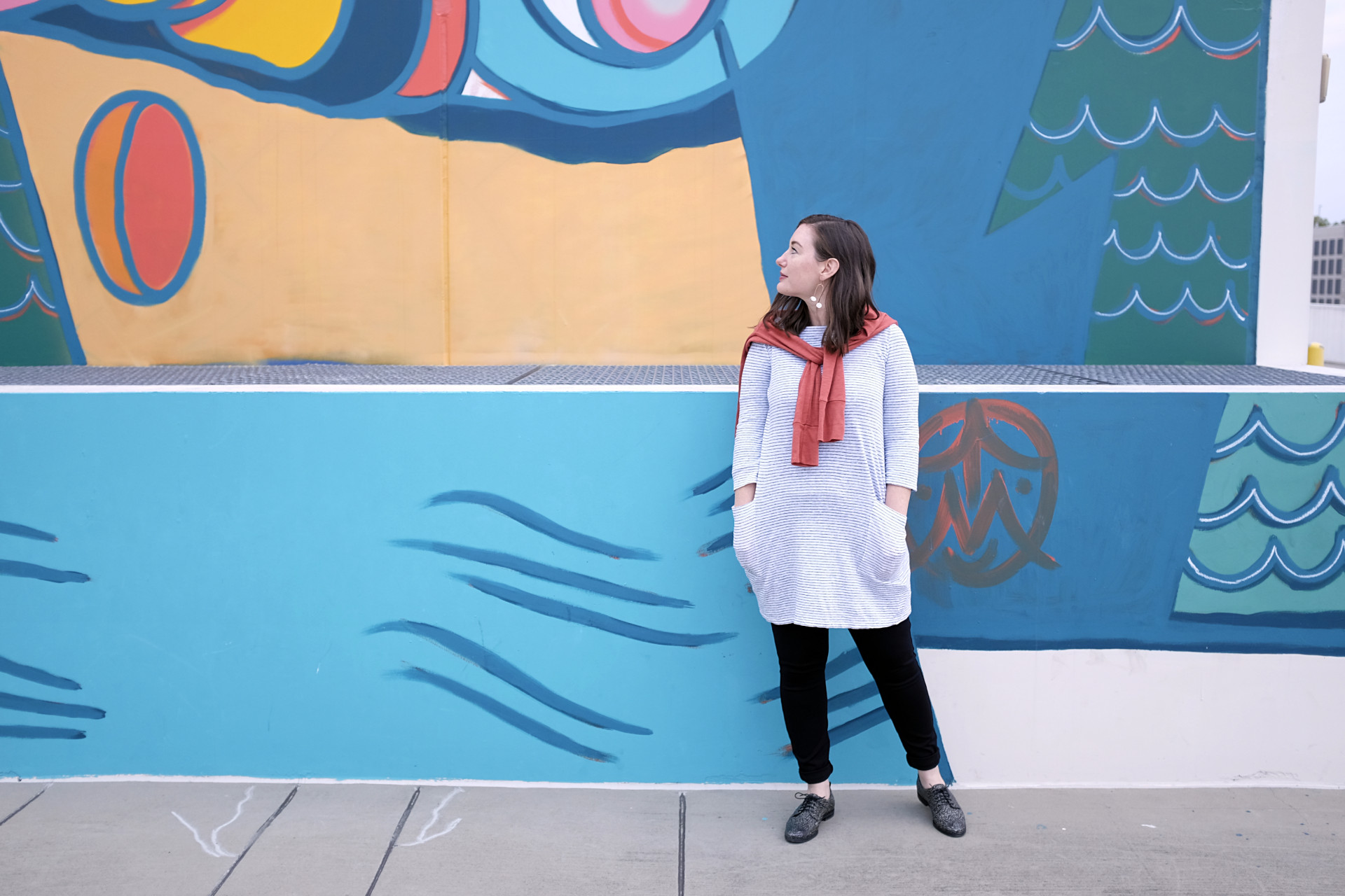 A white woman stands in front of a colorful mural wearing a striped tunic, black pants, black and white oxfords, and a red-orange top is draped around her shoulders. She is wearing abstract butterfly earrings, and they are noticeable because she is looking all the way to her right.