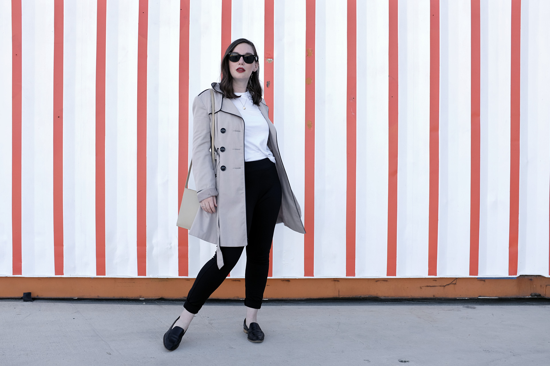 A white woman stands in front of a red and white striped wall. She is wearing a trench, a white tee, black slim pants, and black loafers. Her accessories are minimal jewelry, a crossbody bag, and sunglasses. She is looking at the camera.