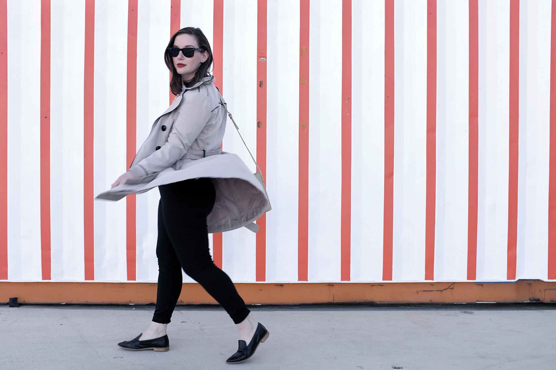 A white woman stands in front of a red and white striped wall. She is wearing a trench, a white tee, black slim pants, and black loafers. Her accessories are minimal jewelry, a crossbody bag, and sunglasses. She is turned to her right but looking at the camera. Her trench coat billows with the motion.