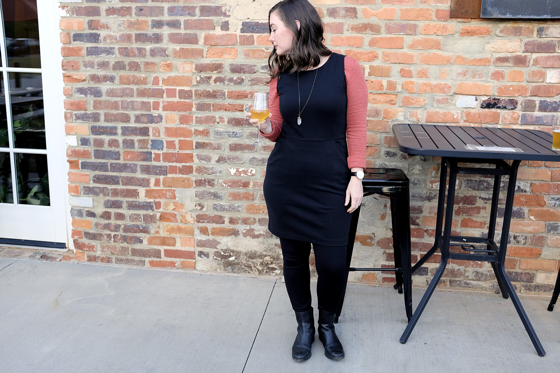 A white woman stands in front of a table which is in front of a brick wall. She is wearing a red-orange long sleeve tee under a black sheath dress, over black ponte pants with chelsea boots. She is holding a beer in a stemmed glass and looking to her right.