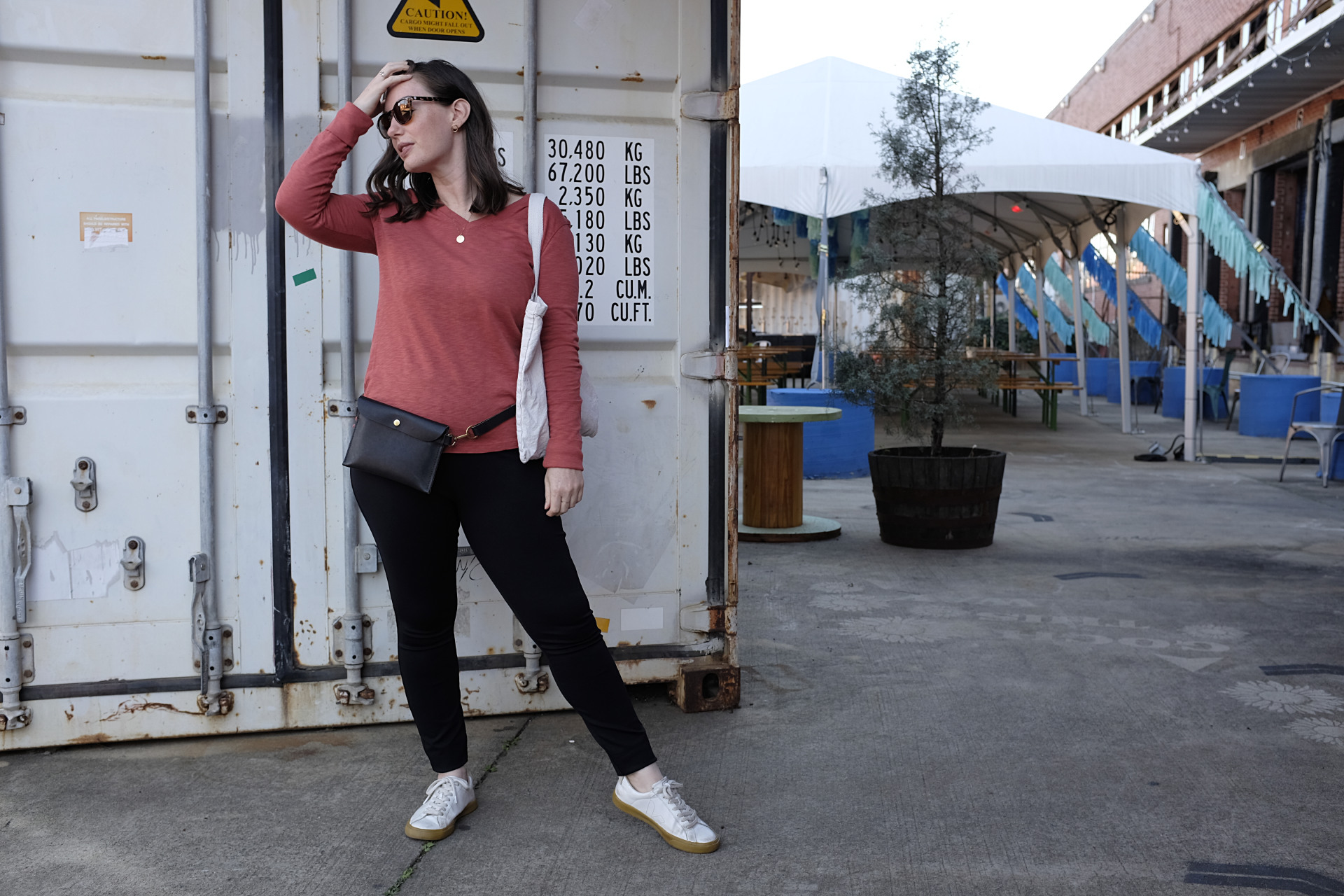 A white woman stands in front of a shipping container, which is in front of a large tent. She is wearing a red-orange long sleeve tee, black pants, white sneakers, a black belt bag, tortiseshell sunglasses, and is carrying a tote. She has her hand in her hair and is starting to her right.