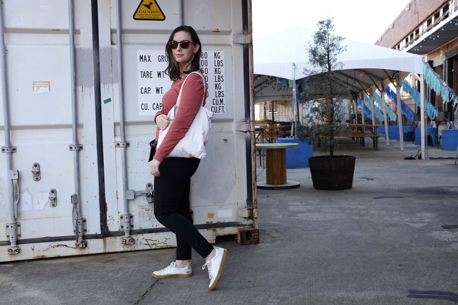 A white woman stands in front of a shipping container, which is in front of a large tent. She is wearing a red-orange long sleeve tee, black pants, white sneakers, a black belt bag, tortiseshell sunglasses, and is carrying a tote. She is facing the camera but her body is pointed to the left.