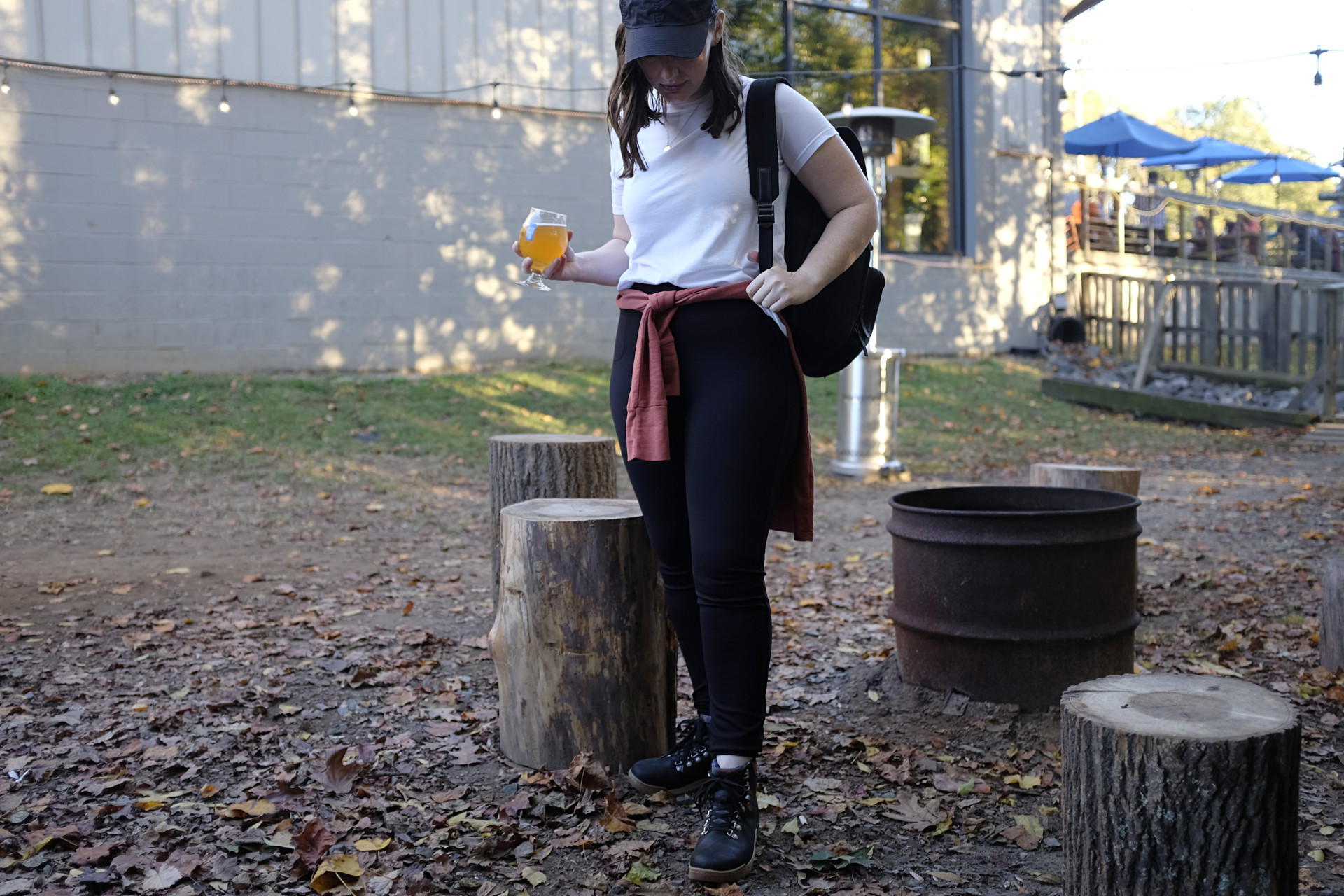 A white woman sits on a stump next to a fire pit. She is wearing a white tee, black pants, a baseball cap, and hiking boots. An orange-red shirt is tied around her waist and she is holding a beer. Her backpack is set on the ground on her right side.