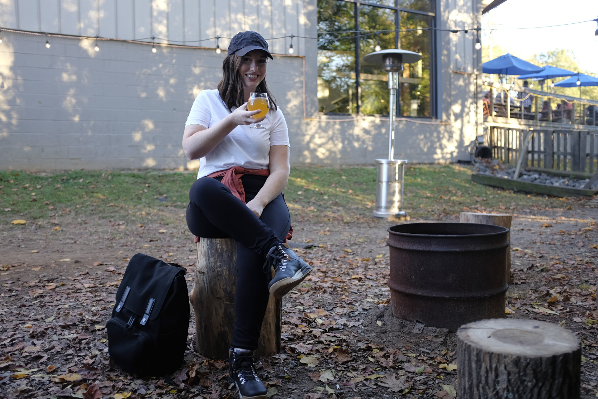 A white woman sits on a stump next to a fire pit. She is wearing a white tee, black pants, a baseball cap, and hiking boots. An orange-red shirt is tied around her waist and she is holding a beer. Her backpack is set on the ground on her right side.