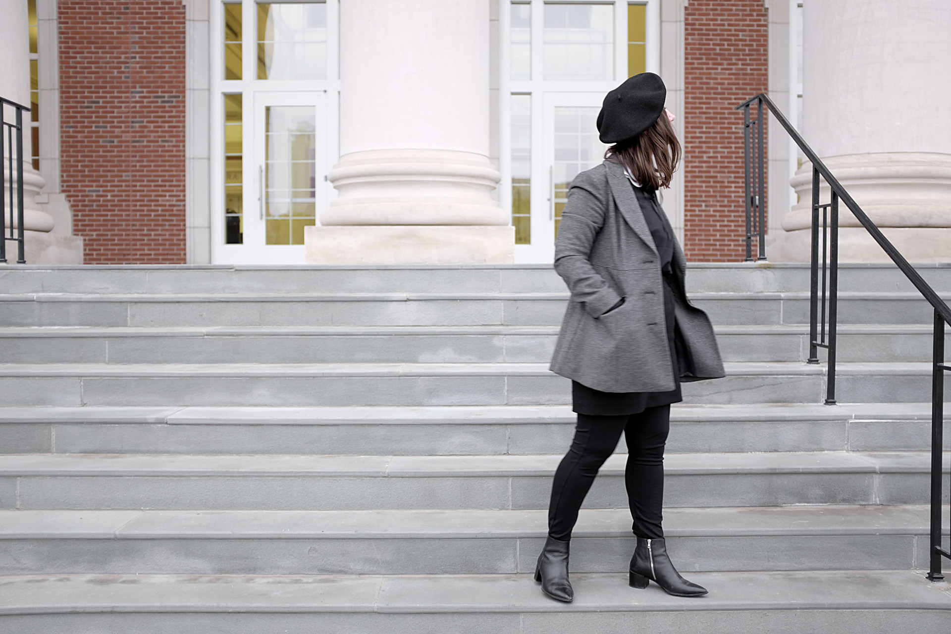A white woman stands on steps with columns in the background. She is wearing a black beret, a white collared shirt under a black sweatshirt, a black wool skirt, a pair of black pants, black boots, and a grey coat.