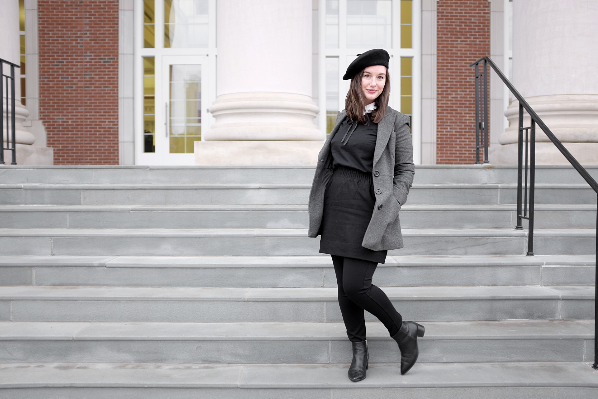 A white woman stands on steps with columns in the background. She is wearing a black beret, a white collared shirt under a black sweatshirt, a black wool skirt, a pair of black pants, black boots, and a grey coat.