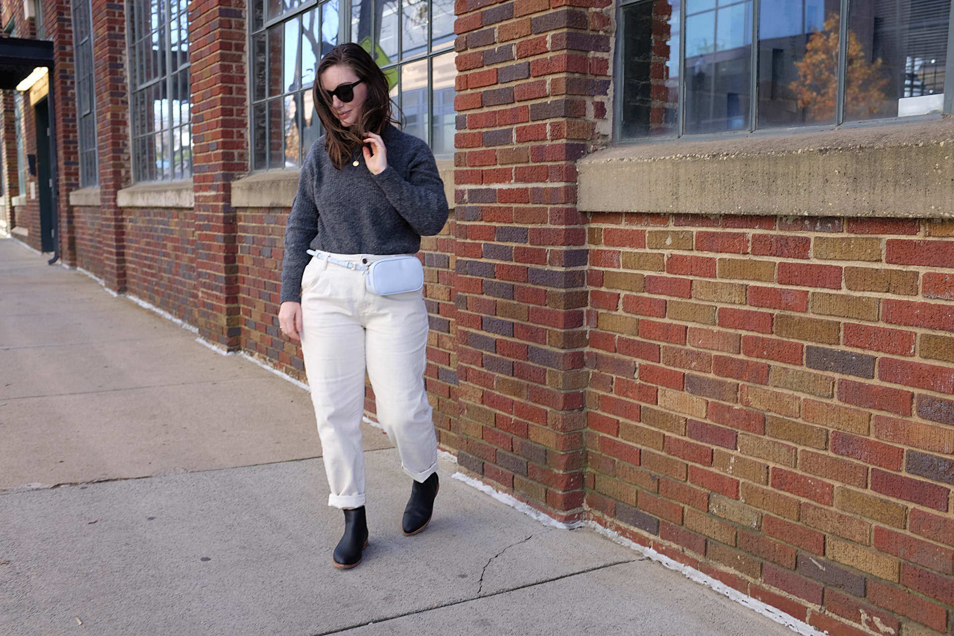 Alyssa is standing in front of a brick building. She is wearing a grey sweater with off-white pants, black sunglasses, a light blue belt bag, and black boots. She has her left hand in her hair and it is windy.