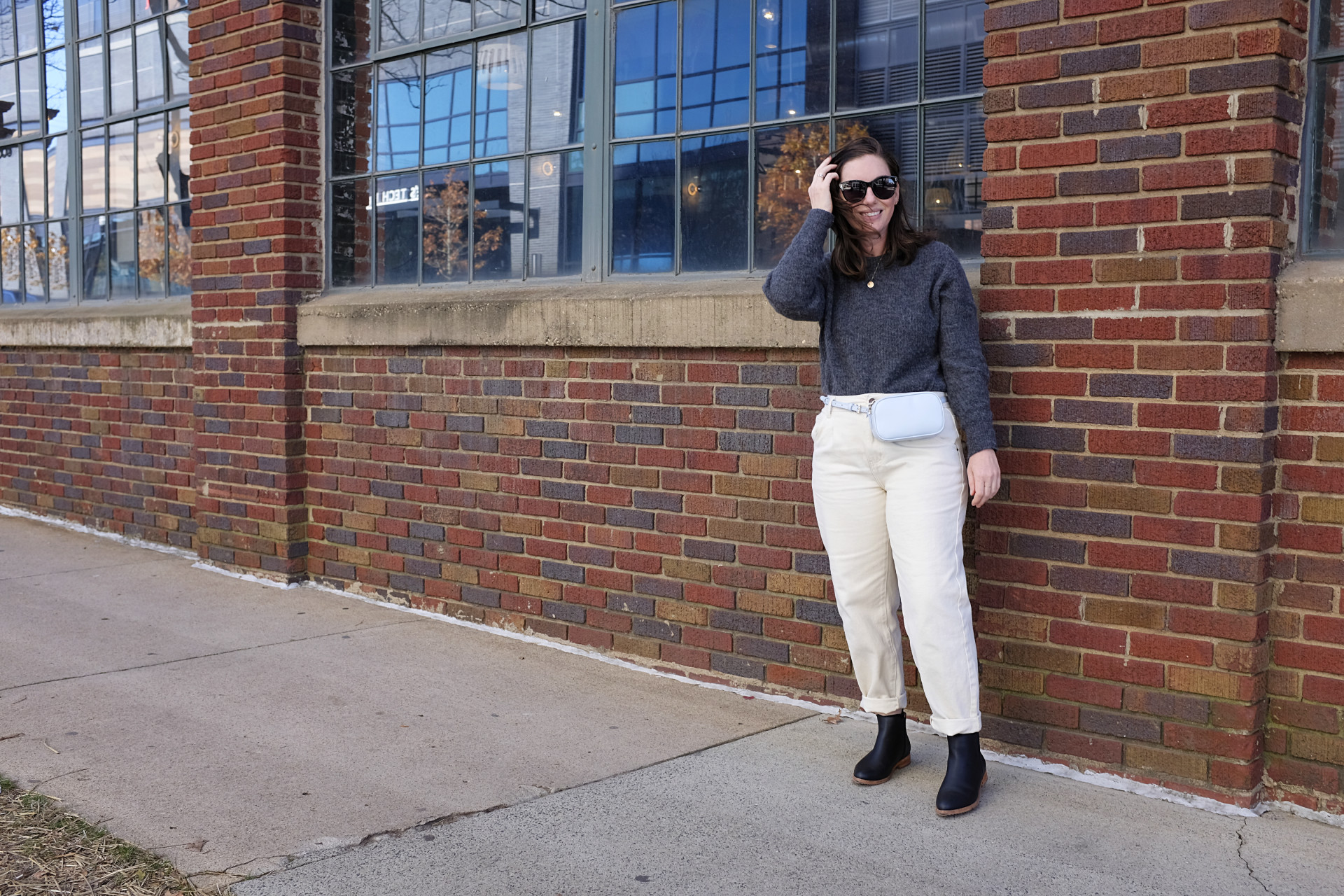 Alyssa is standing in front of a brick building. She is wearing a grey sweater with off-white pants, black sunglasses, a light blue belt bag, and black boots. She has her right hand in her hair and it is windy.