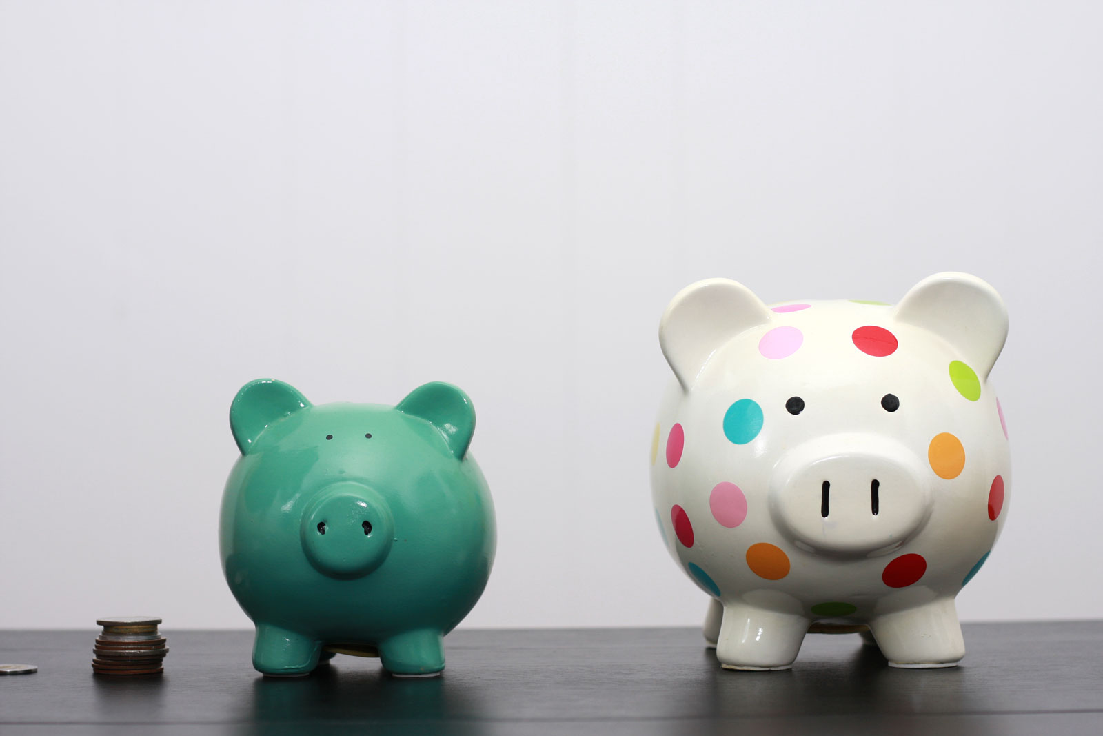 Two piggy banks, one is teal and one is white with polka dots. There is a stack of coins on the left.