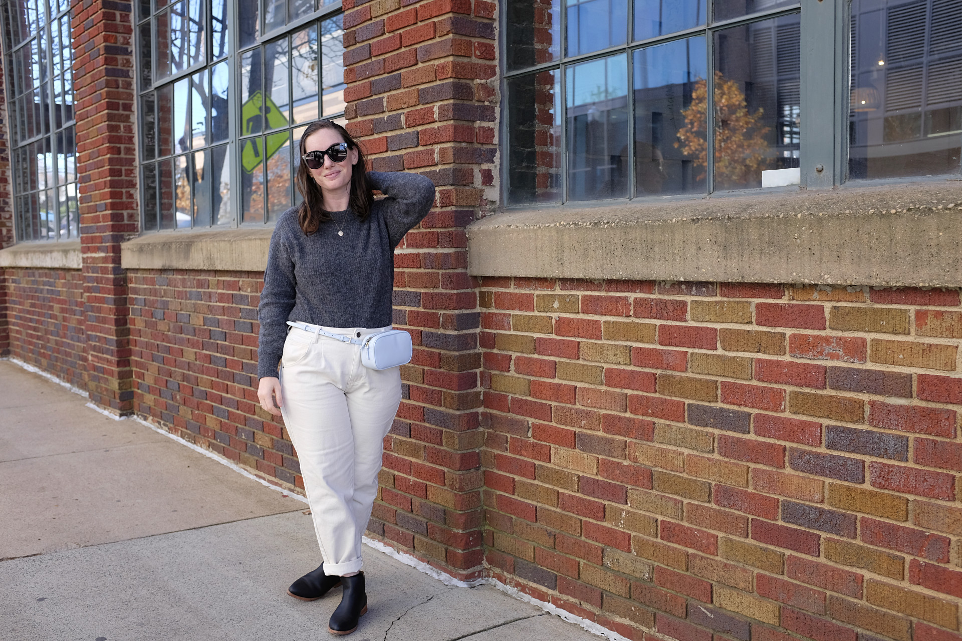 Alyssa is standing in front of a brick building. She is wearing a grey sweater with off-white pants, black sunglasses, a light blue belt bag, and black boots. She has her left hand in her hair and it is windy.