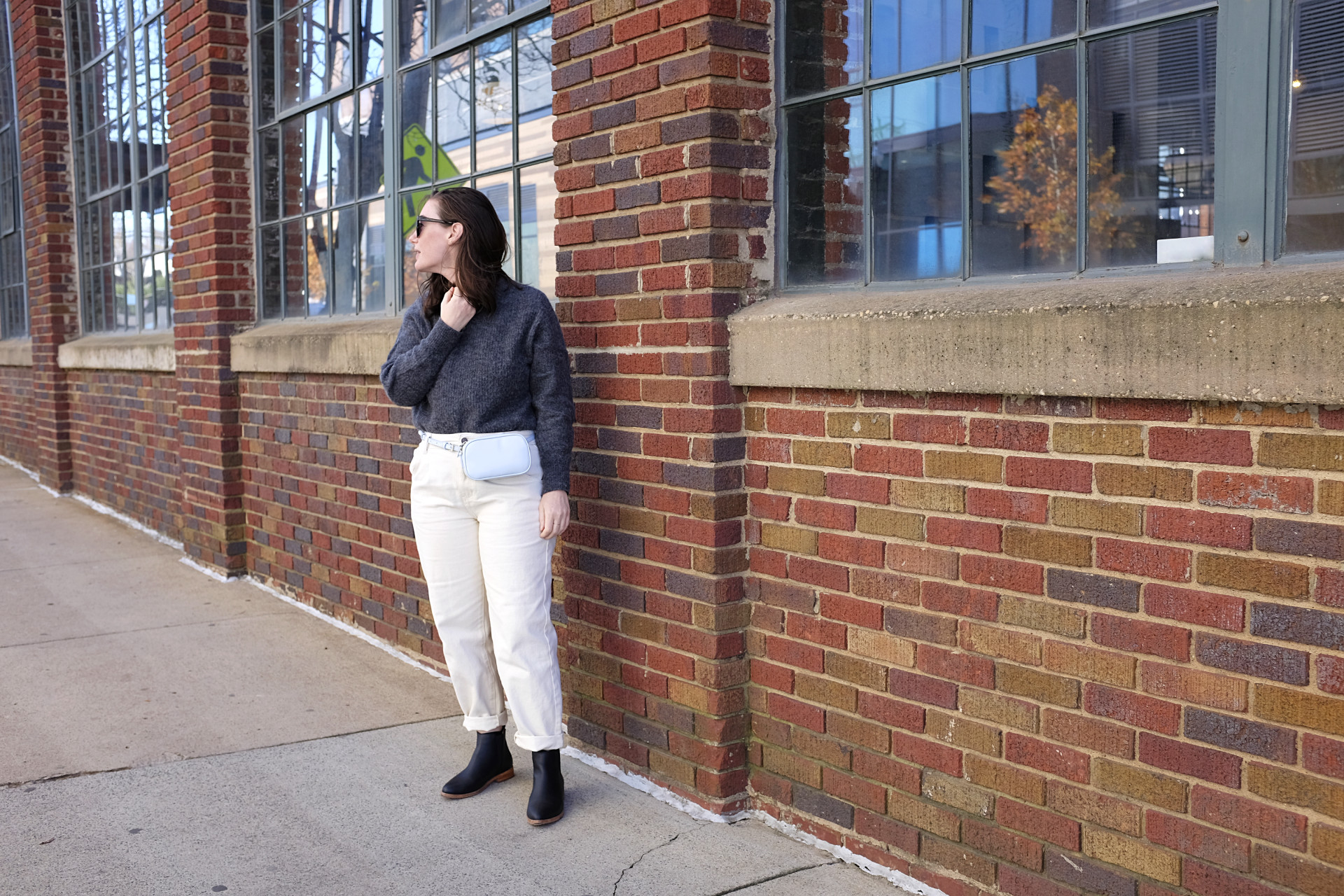 Alyssa is standing in front of a brick building. She is wearing a grey sweater with off-white pants, black sunglasses, a light blue belt bag, and black boots. She is looking off to her right.