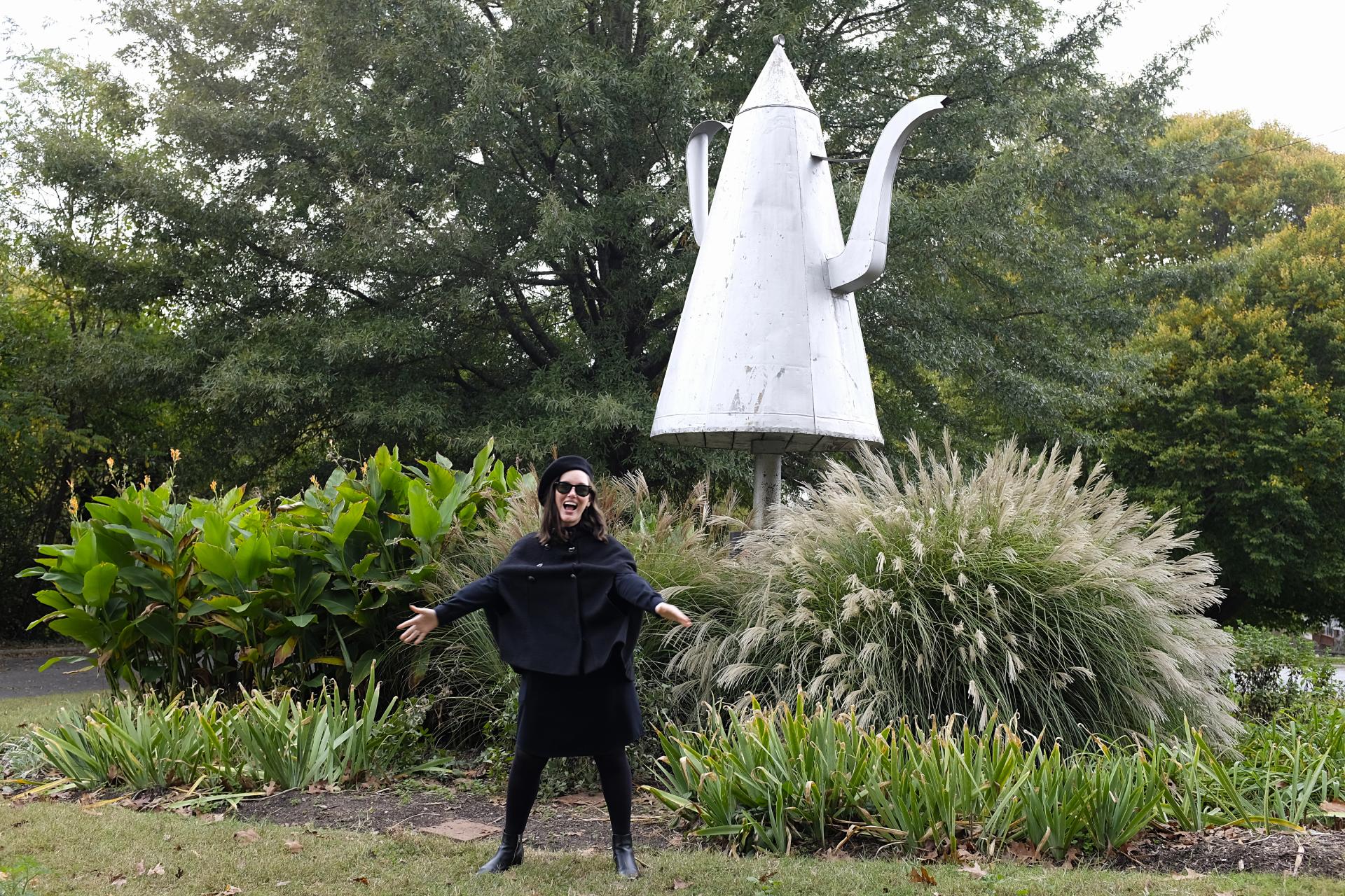 Alyssa stands in front of a large coffee pot sculpture and makes a silly face/gesture