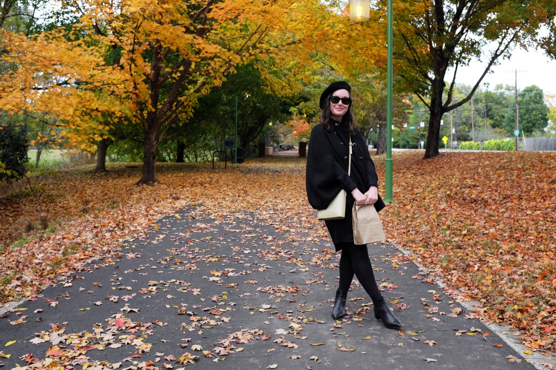 Alyssa stands on a leaf strewn path. She is wearing a black dress, cape, beret, and tights. She is holding a paper sack.