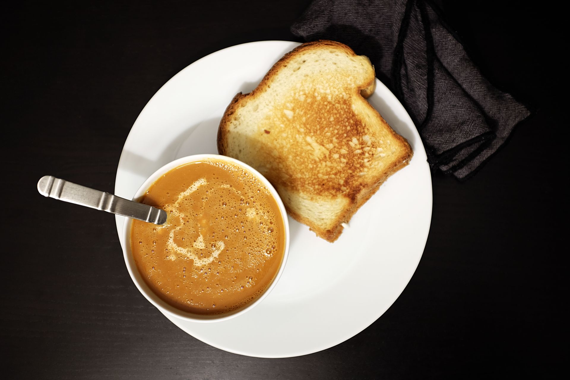 A white plate with a bowl of tomato soup and a grilled cheese sandwich