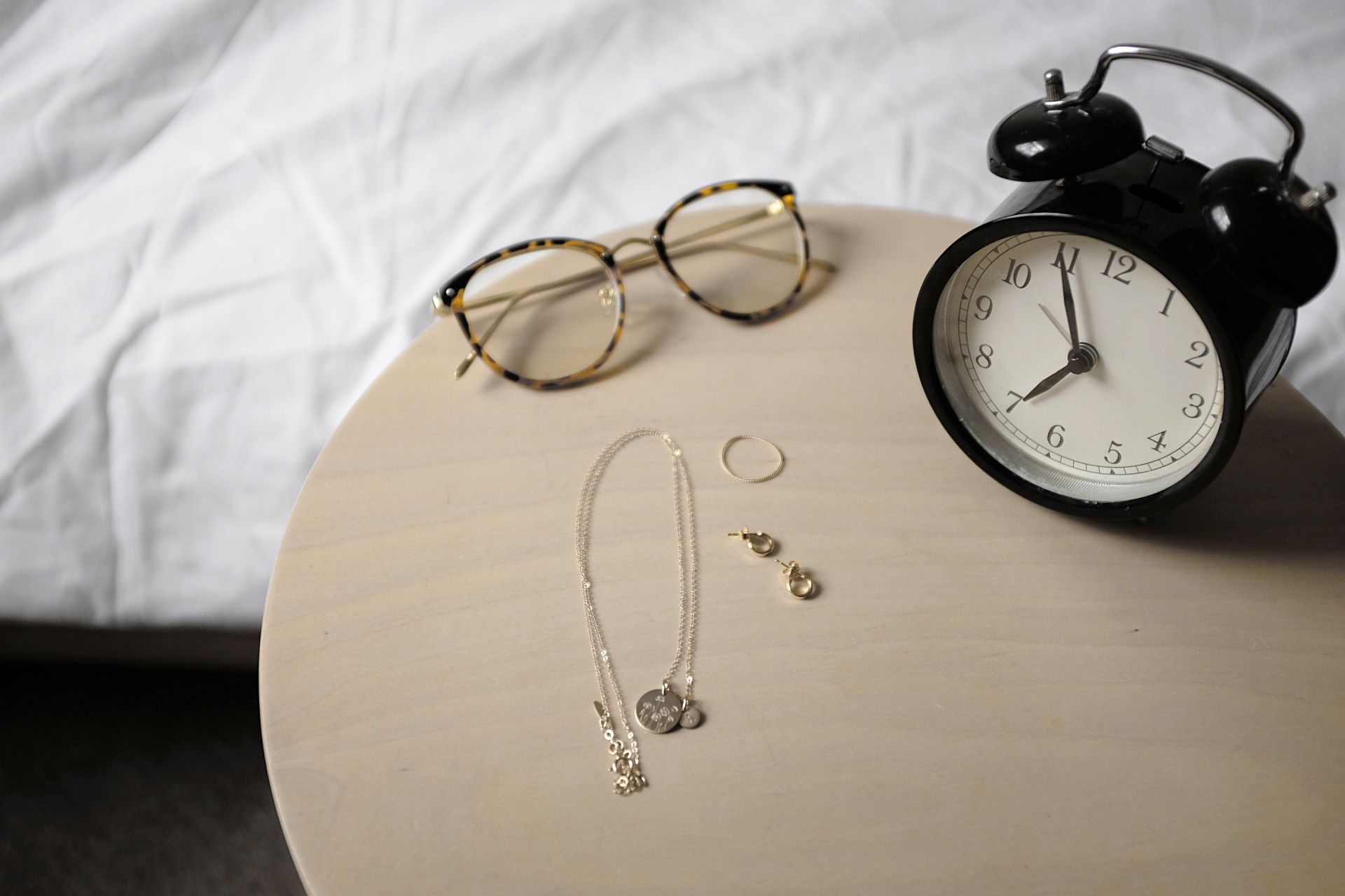 A pair of glasses, a clock, and a necklace, ring, and earrings sit on a nightstand