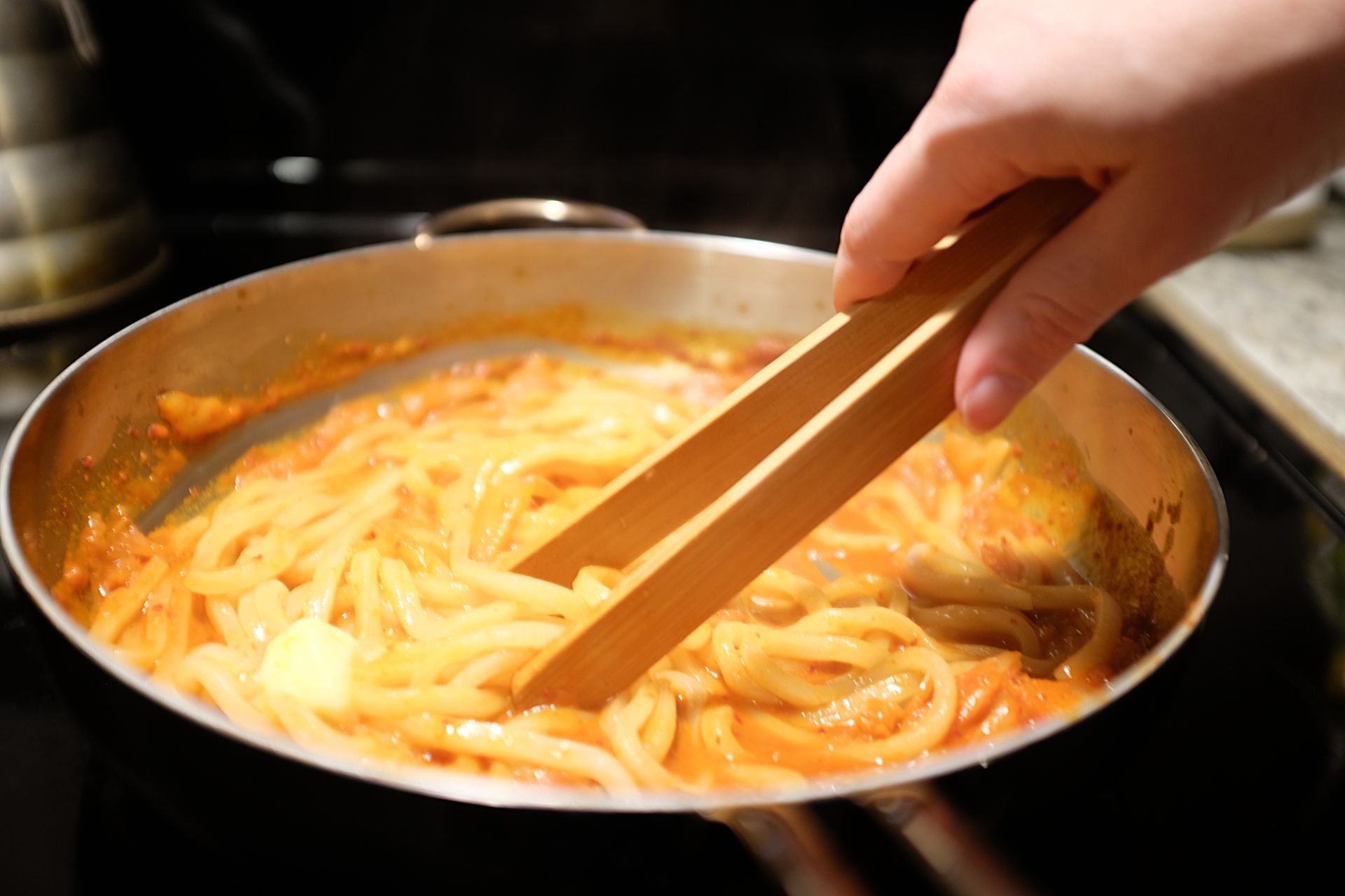A white woman's hand stirs udon noodles in a reddish sauce with wooden tongs