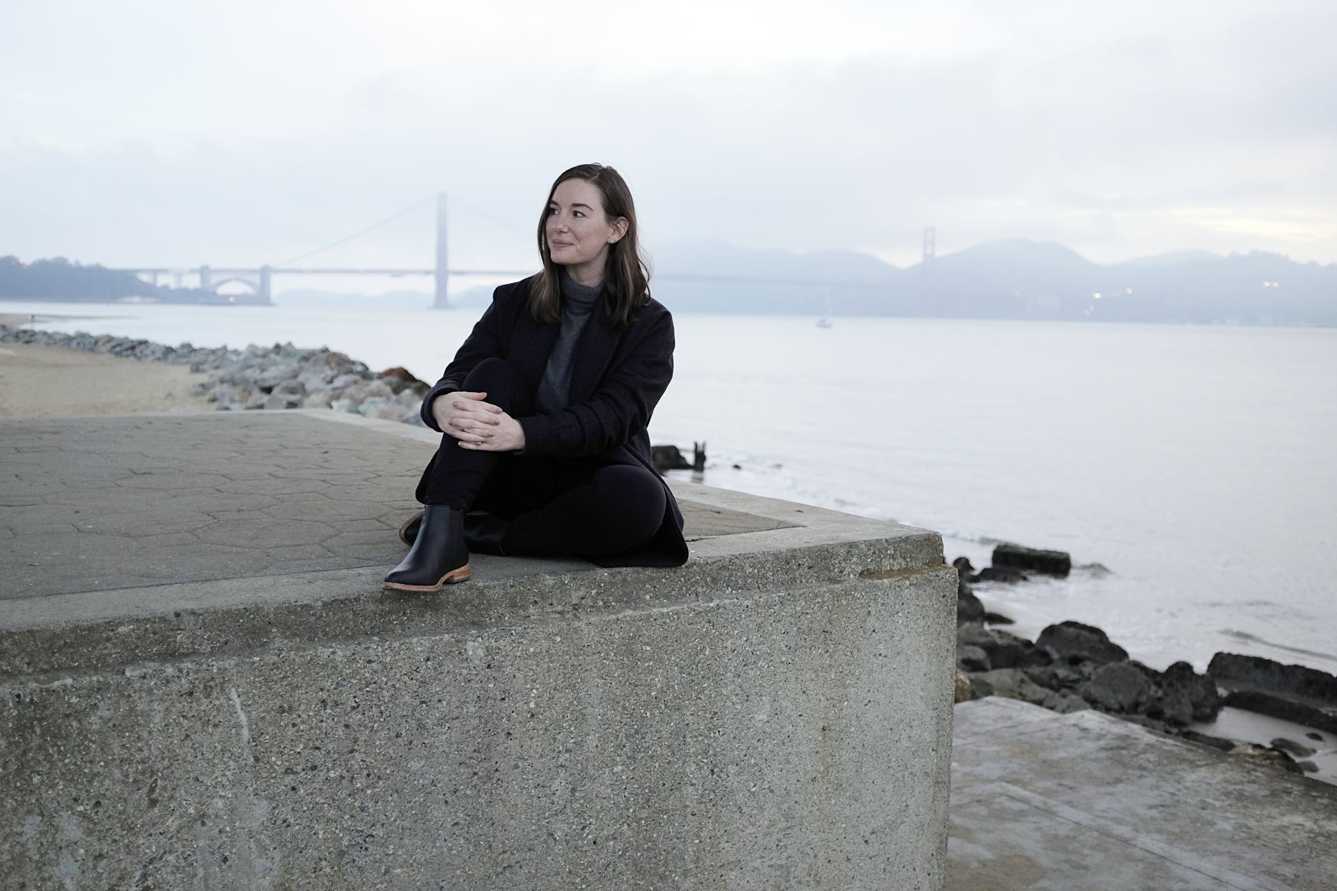 Alyssa is sitting on a concrete wall in front of the Golden Gate bridge. She is wearing black pants, a grey sweater, a grey coat, and black boots.