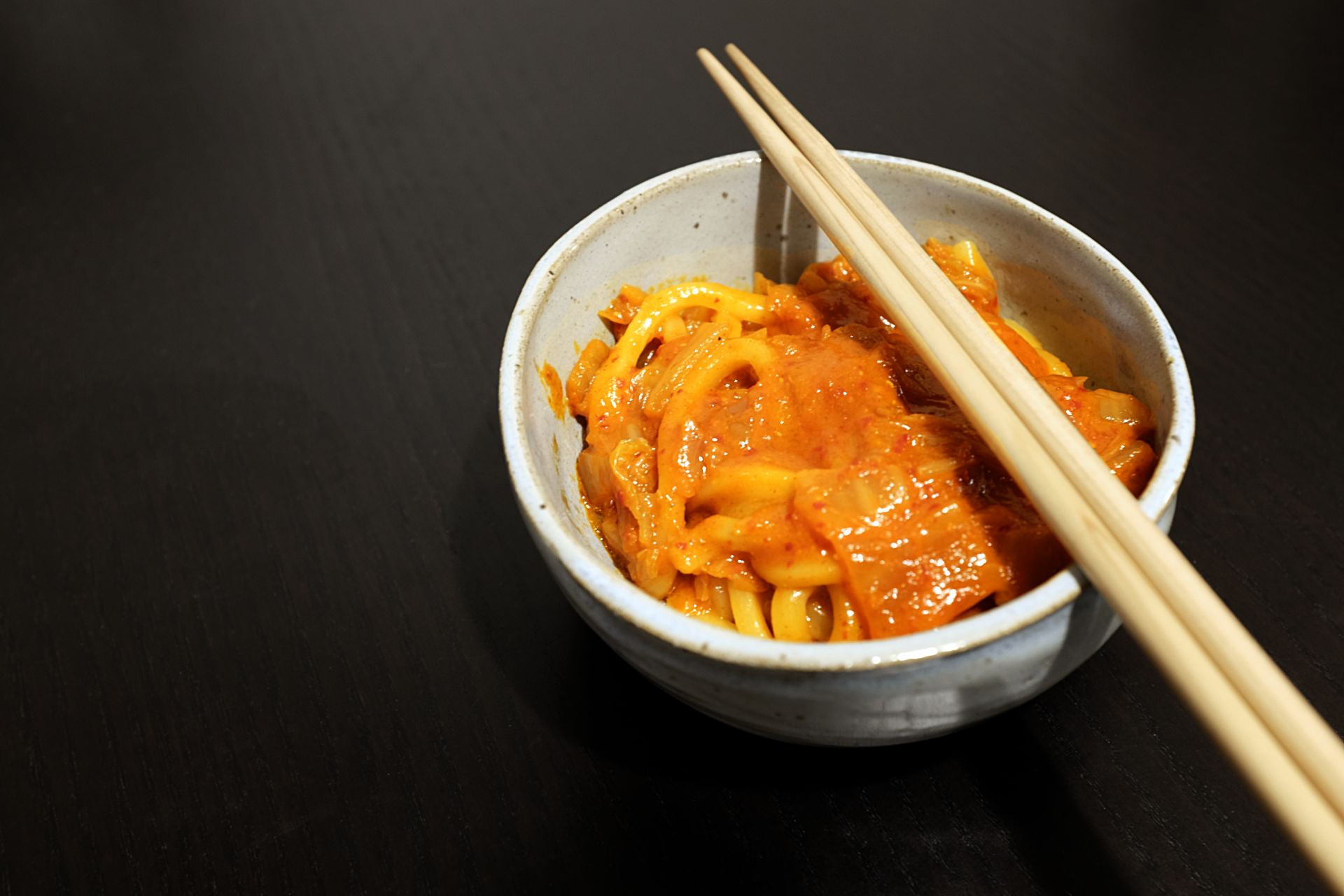 A handmade blue bowl with udon noodles in a reddish sauce. A pair of chopsticks is resting on the bowl