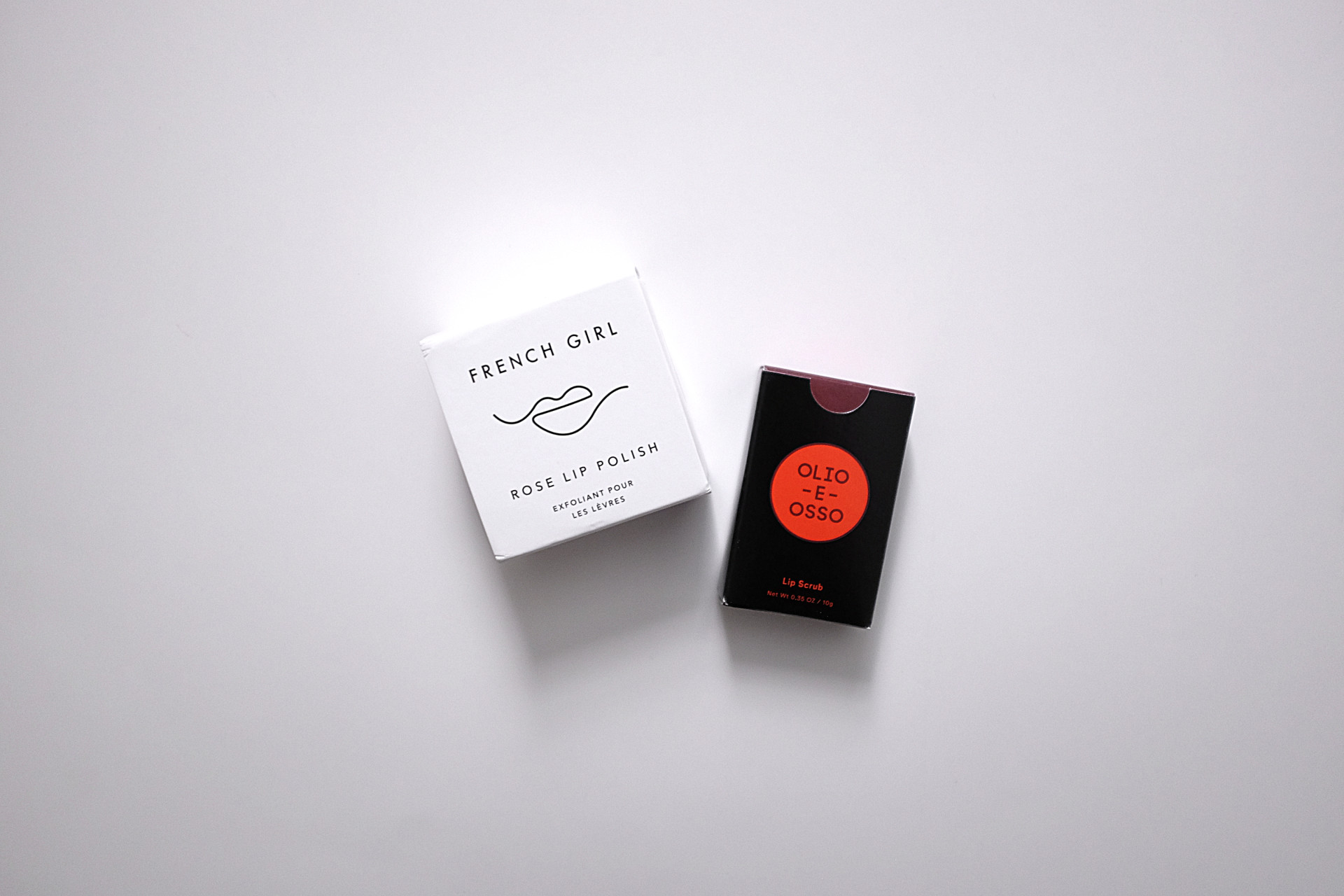 Two small boxes on a white background. On the left is a white box that reads "FRENCH GIRL rose lip polish" with an abstract lip line drawing. On the right is a black box with a red circle which has "Olio e Osso" printed in it