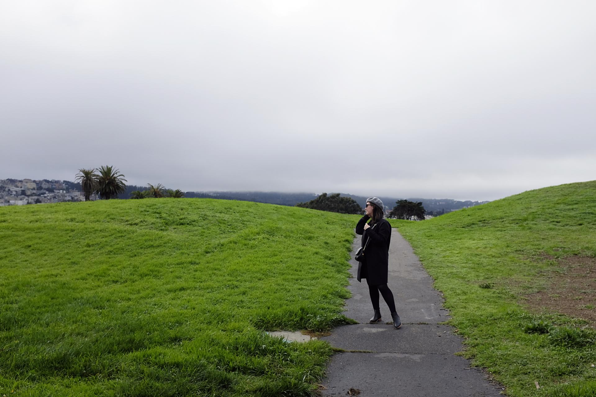 Alyssa stands in a green field in San Francisco, wearing all black and grey, black boots, and a beret. She is looking to her right