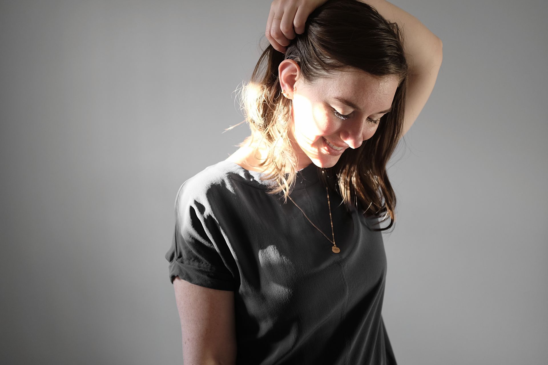 Alyssa stands in a beam of sunshine, wearing a navy silk short sleeve dress, and a pendant necklace with two mini hoops in her right ear. She is pulling her hair out of the way and smiling downward