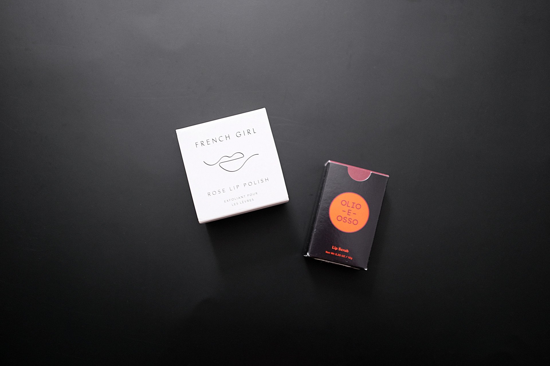 Two small boxes on a black background. On the left is a white box that reads "FRENCH GIRL rose lip polish" with an abstract lip line drawing. On the right is a black box with a red circle which has "Olio e Osso" printed in it