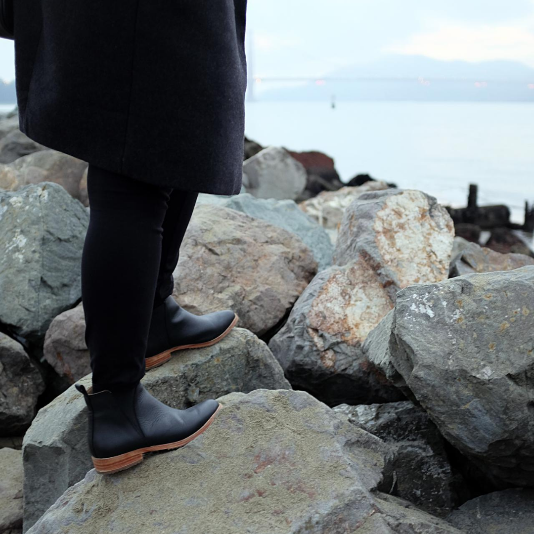 Alyssa is standing on rocks wearing black pants, black boots. It's a close up shot of her calves and feet, and the Golden Gate Bridge is visible in the background.