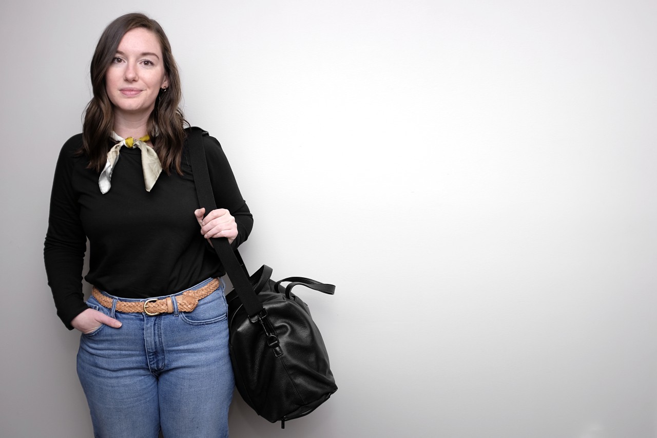 Alyssa stands in the left of the frame, wearing a black sweatshirt, blue jeans, tan belt, and a green scarf. She is carrying the Everywhere Bag from Away on her left shoulder