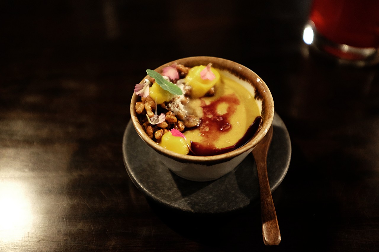A bowl of yellowish custard topped with candied pecans, nasturtiums, and a black glaze