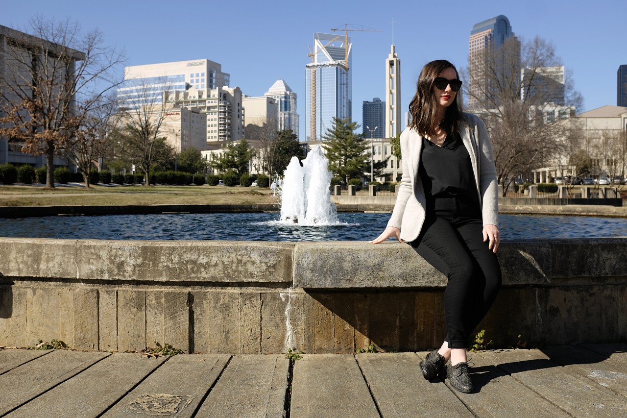 Alyssa sits on the edge of a fountain in front of the Charlotte skyline. She is wearing a black top, black jeans, a grey sweater, black Oxfords, and black sunglasses