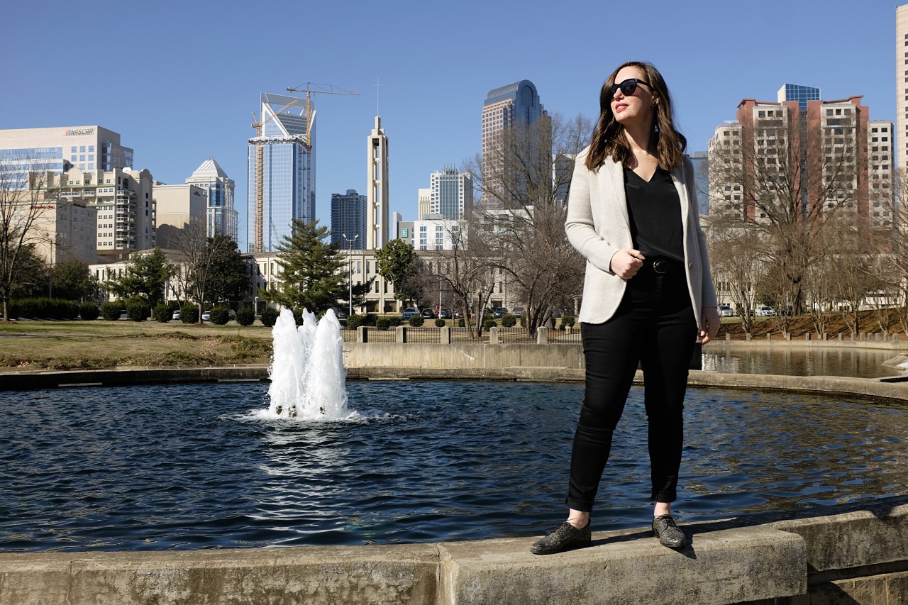 A white woman stands on the ledge of a fountain wearing a black top, black jeans, a grey sweater blazer, and black oxfords. In the background is the Charlotte skyline