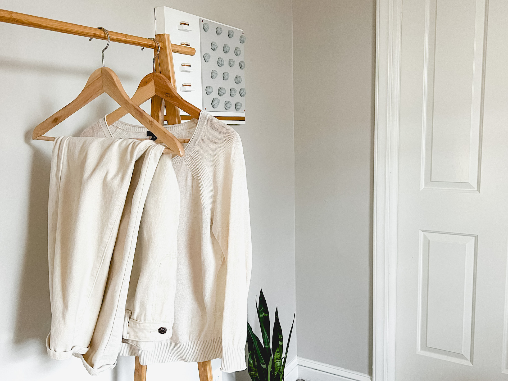 A pair of white jeans and a white sweater hang on a wooden clothes rack. In the background is a piece of fiber art an a snake plant.