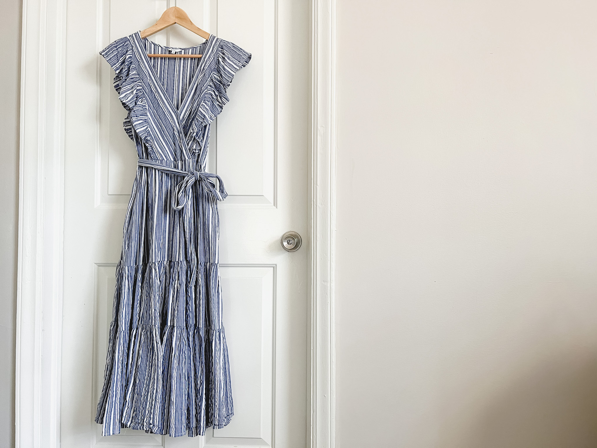 A ruffled blue and white striped dress hangs on a wooden hanger against a white door.