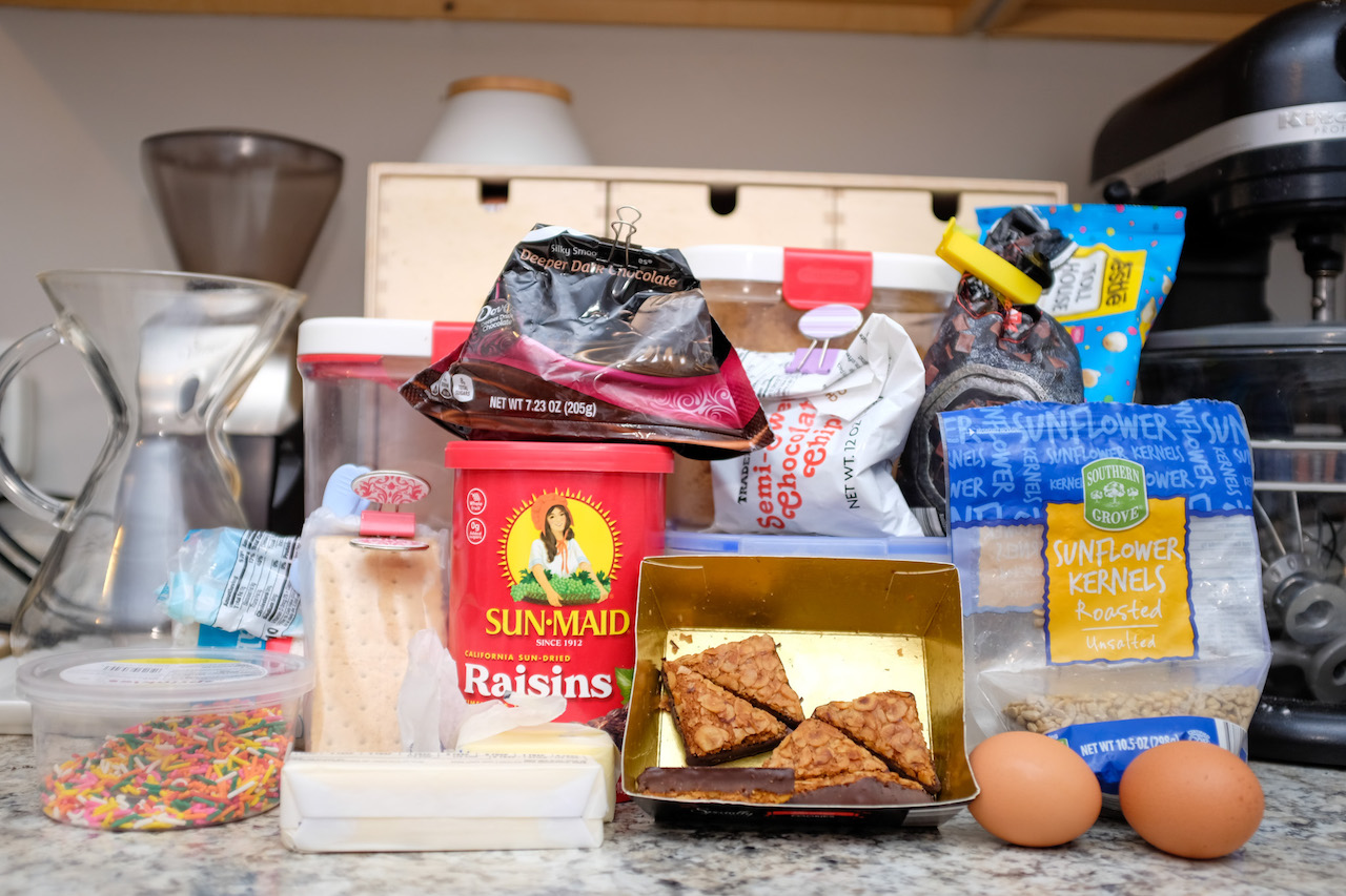 An assortment of cookie ingredients: flour, sugar, eggs, butter, nuts, seeds, dried fruit, etc.