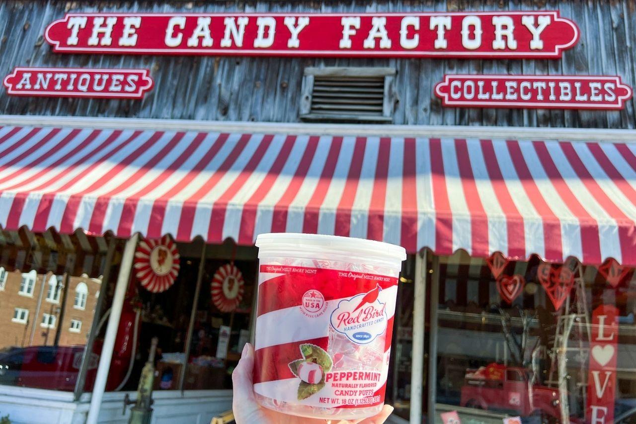 Alyssa's hand holds a tub of Red Bird Candies in front of The Candy Factory storefront