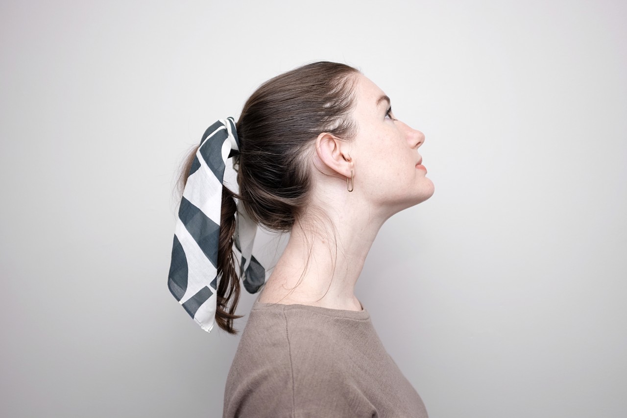 Alyssa is shown in profile, looking up. She is wearing a brown top, brass earrings, and a bandana tied around her ponytail