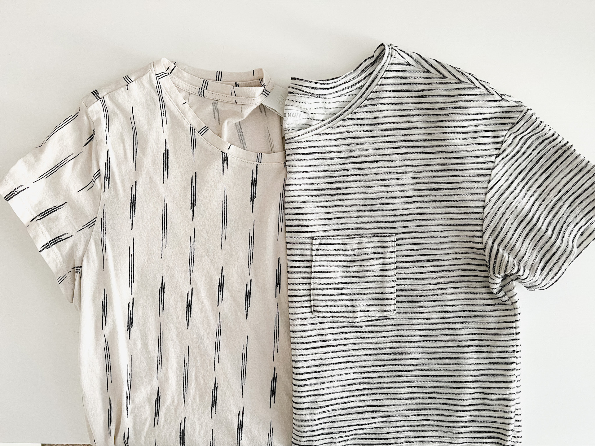 Two patterned t-shirts folded in half and placed next to each other so that they look like one. The left has a cream background and small vertical patches of stripes, and the one on the right is a white t-shirt with thin black horizontal stripes and a pocket at the chest.