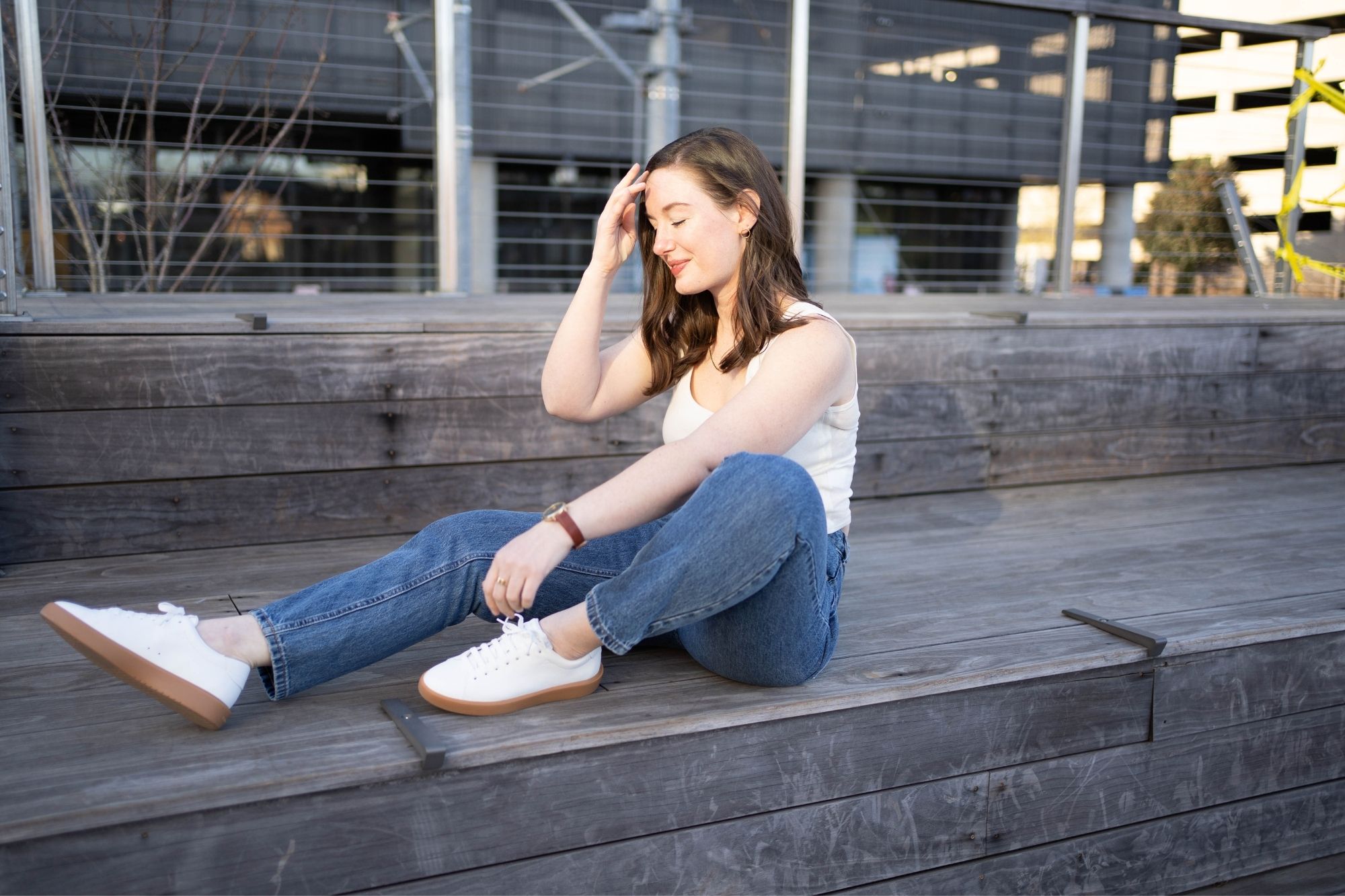 Alyssa wears blue denim, a cream tank, and white sneakers and sits on a wooden step