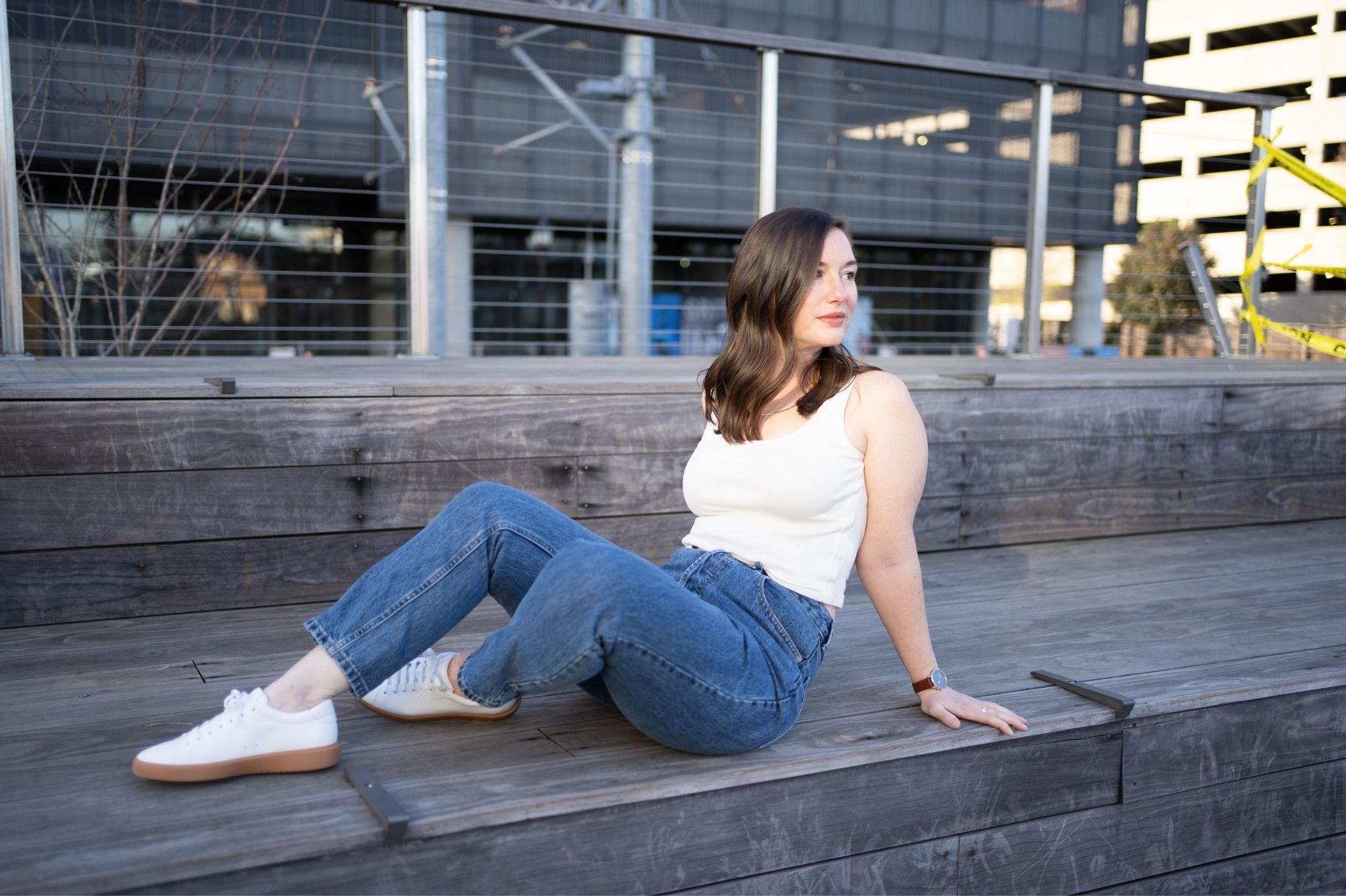 Alyssa wears blue denim, a cream tank, and white sneakers and sits on a wooden step