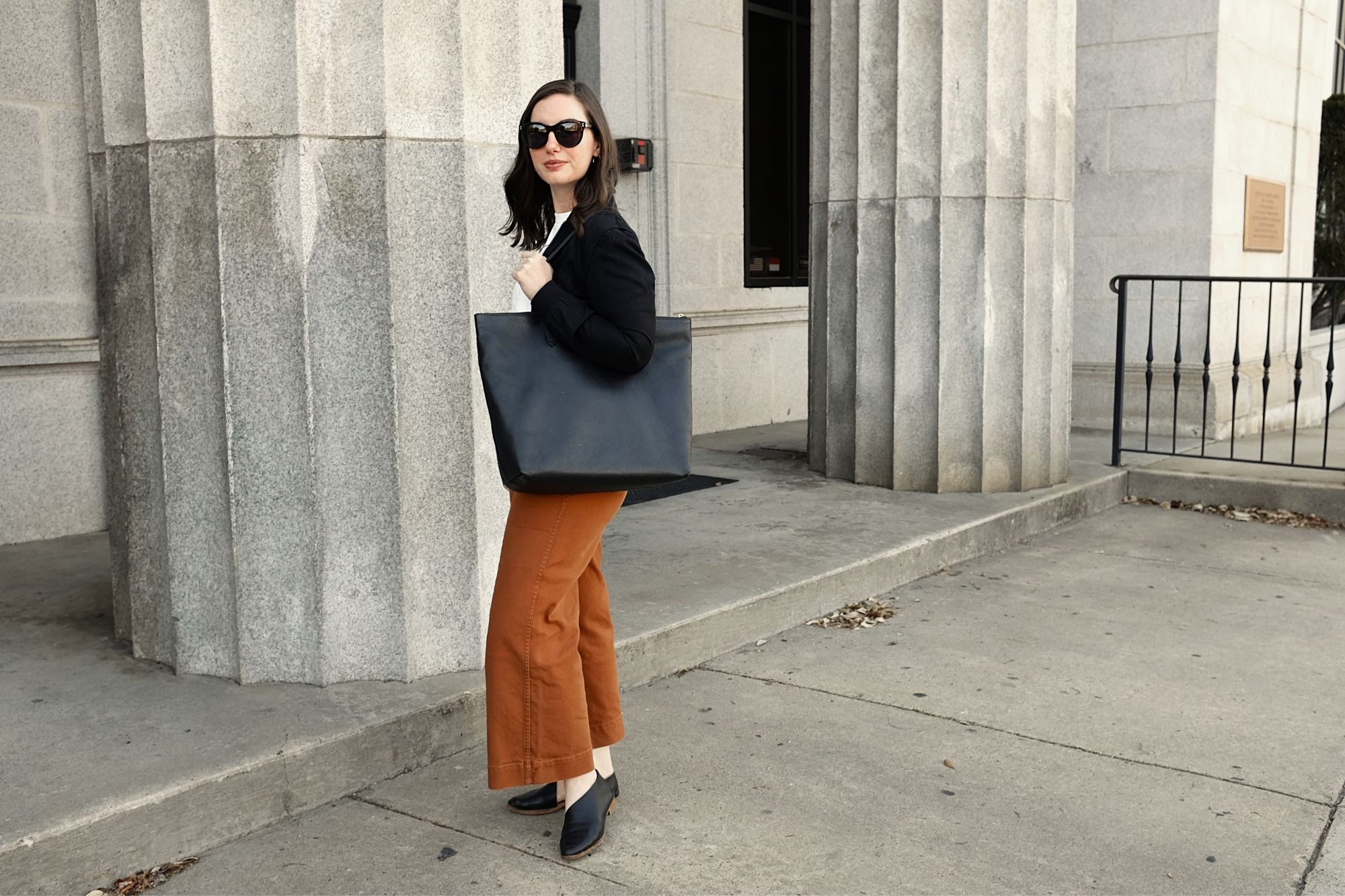 Alyssa wears a black jacket and brown pants with the tote