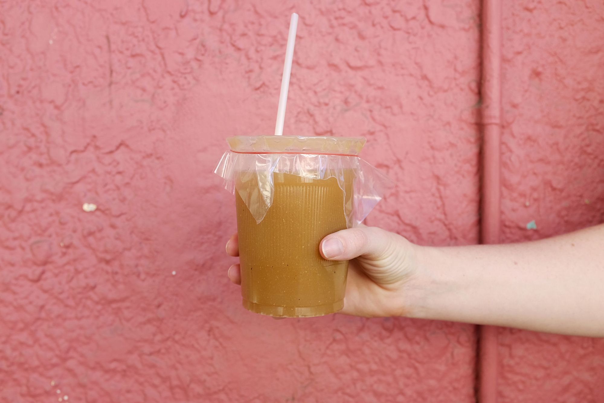 A hand holds a cup of Tejuino in front of a pink wall
