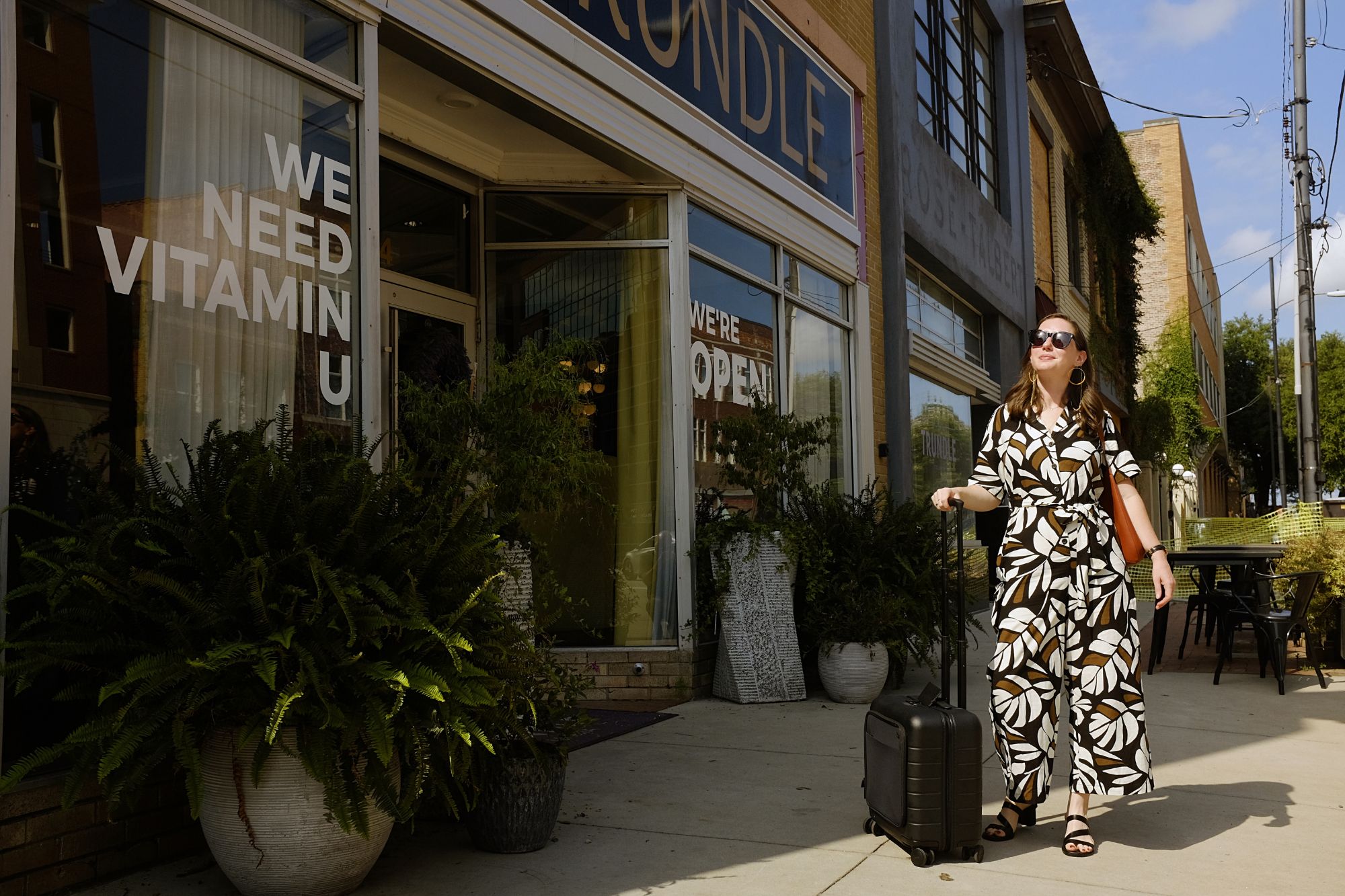 Alyssa stands in front of Hotel Trundle with a suitcase. She is wearing a botanical pattern jumpsuit