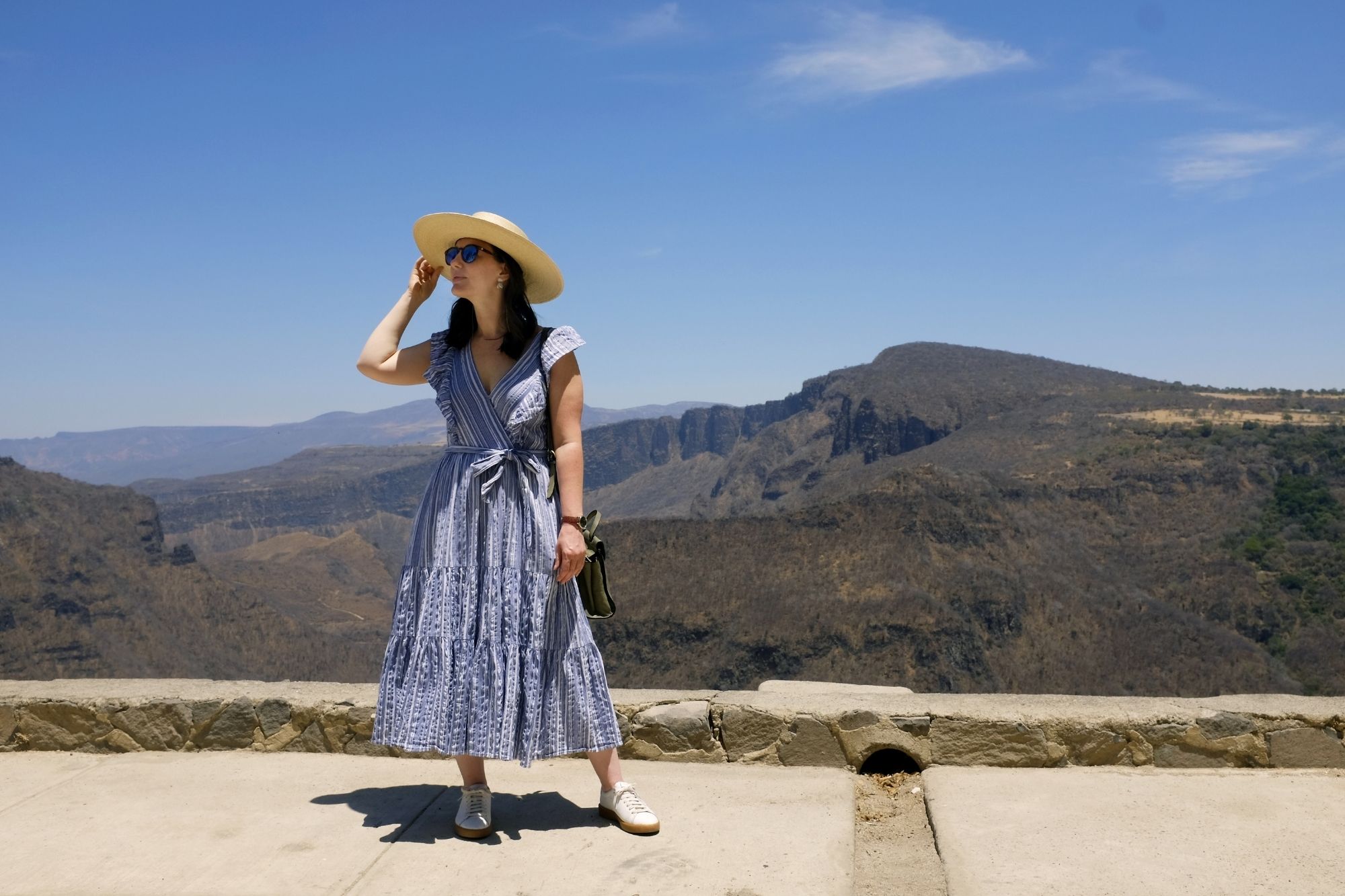 Alyssa looks to the right, she is standing in front of a canyon wearing a blue dress and a straw hat