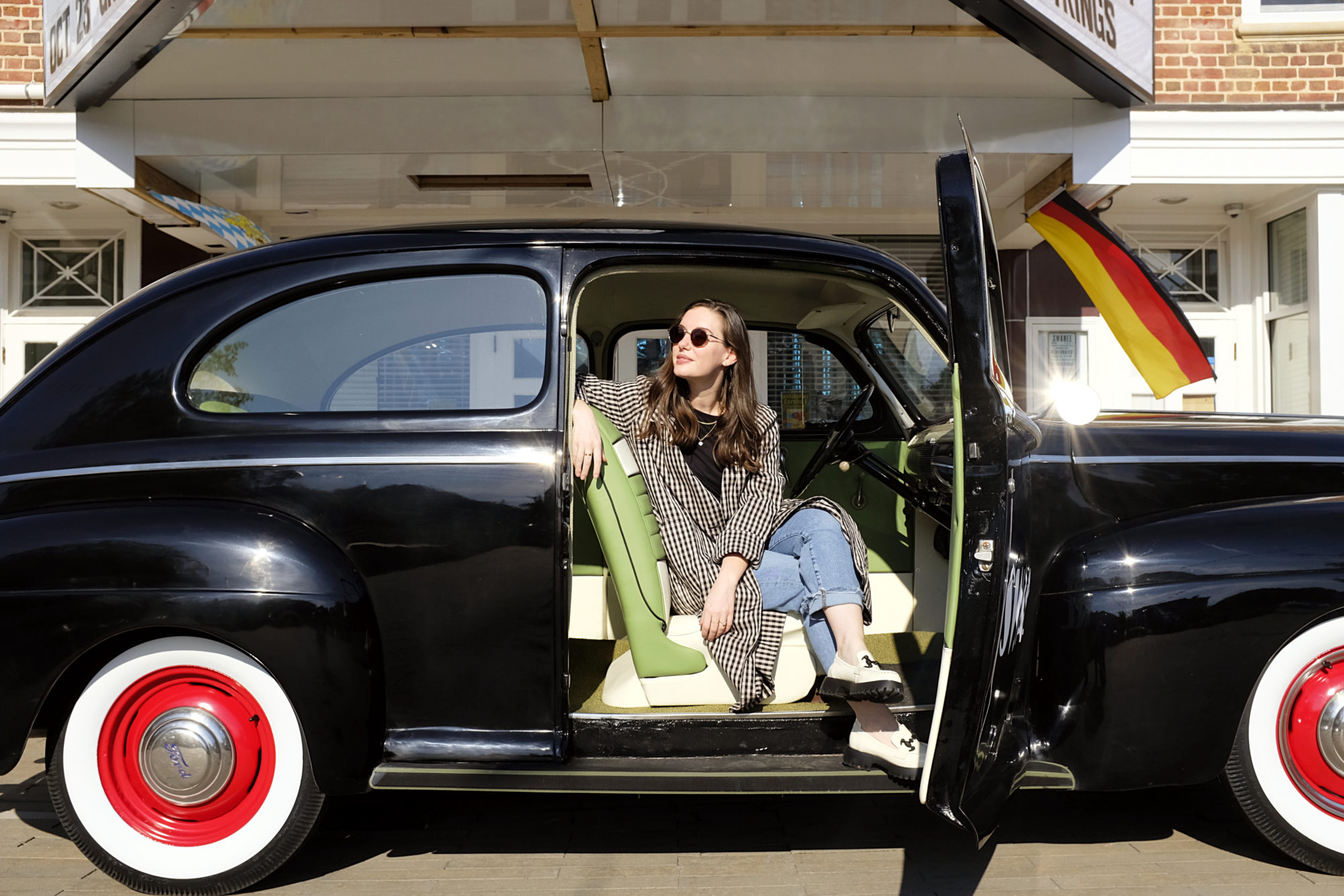 Alyssa sits in a vintage car wearing a black top, blue jeans, white loafers, and gingham trench