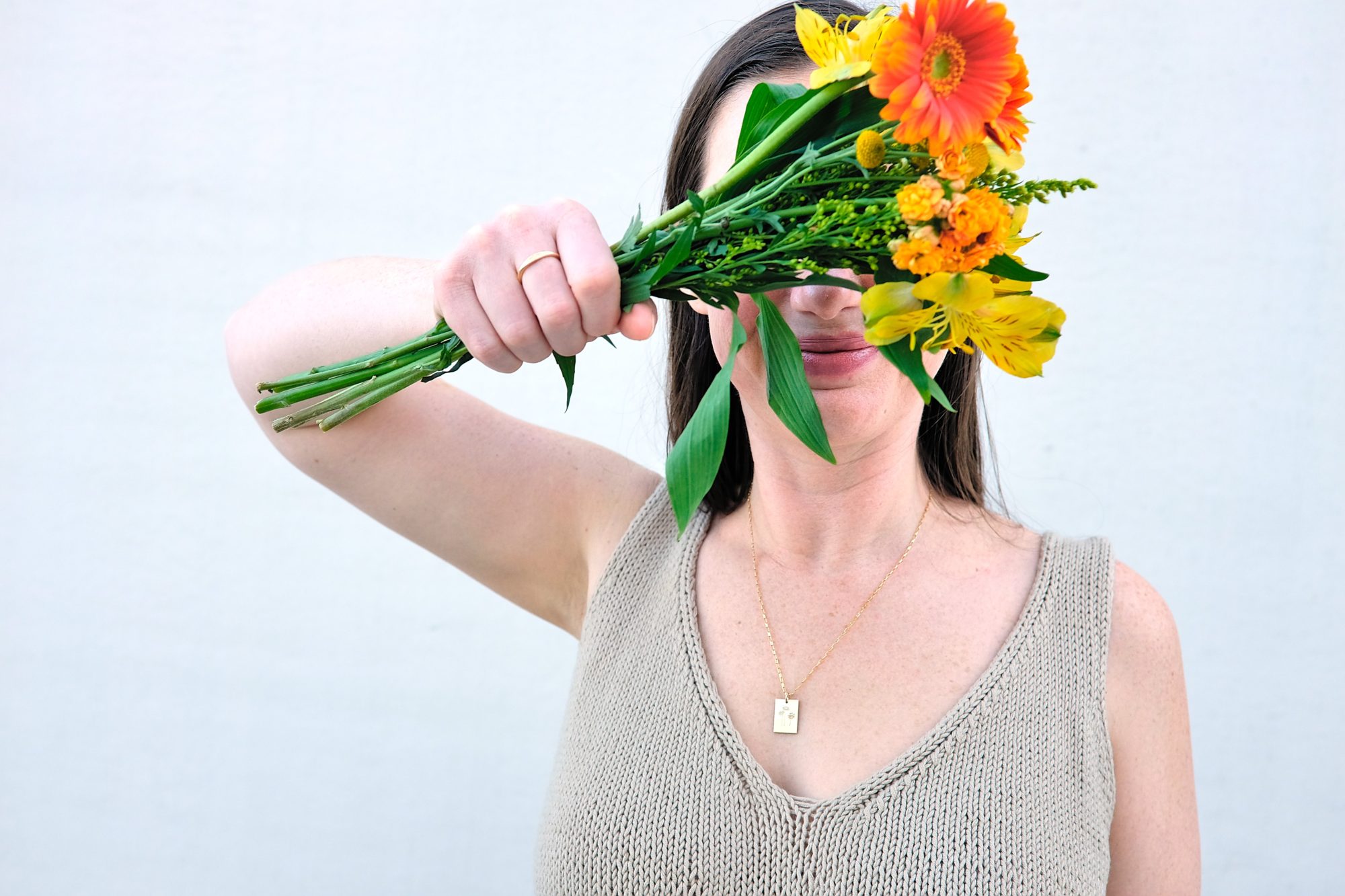 Alyssa holds a bouquet playfully in front of her face while wearing two pieces of jewelry from GLDN