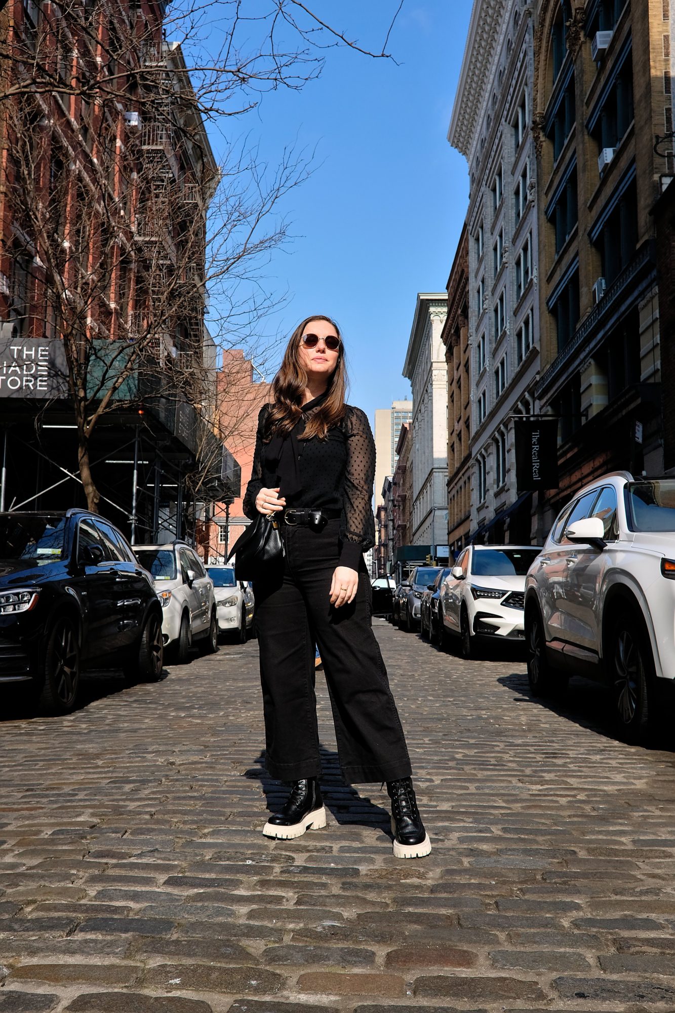 Alyssa stands on a street in SoHo in a Swiss dot top, black pants, and black boots