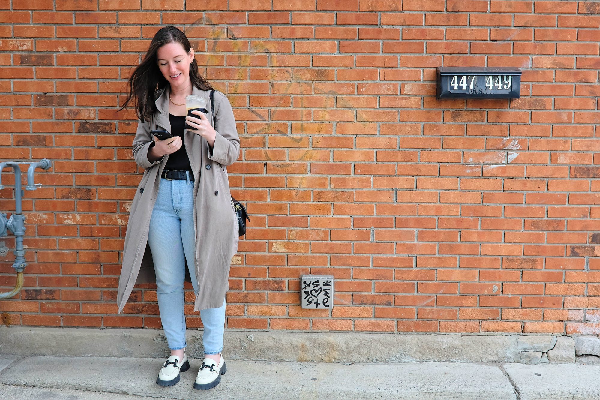 Alyssa stands on a sidewalk in Detroit, ordering an Uber and drinking a coffee
