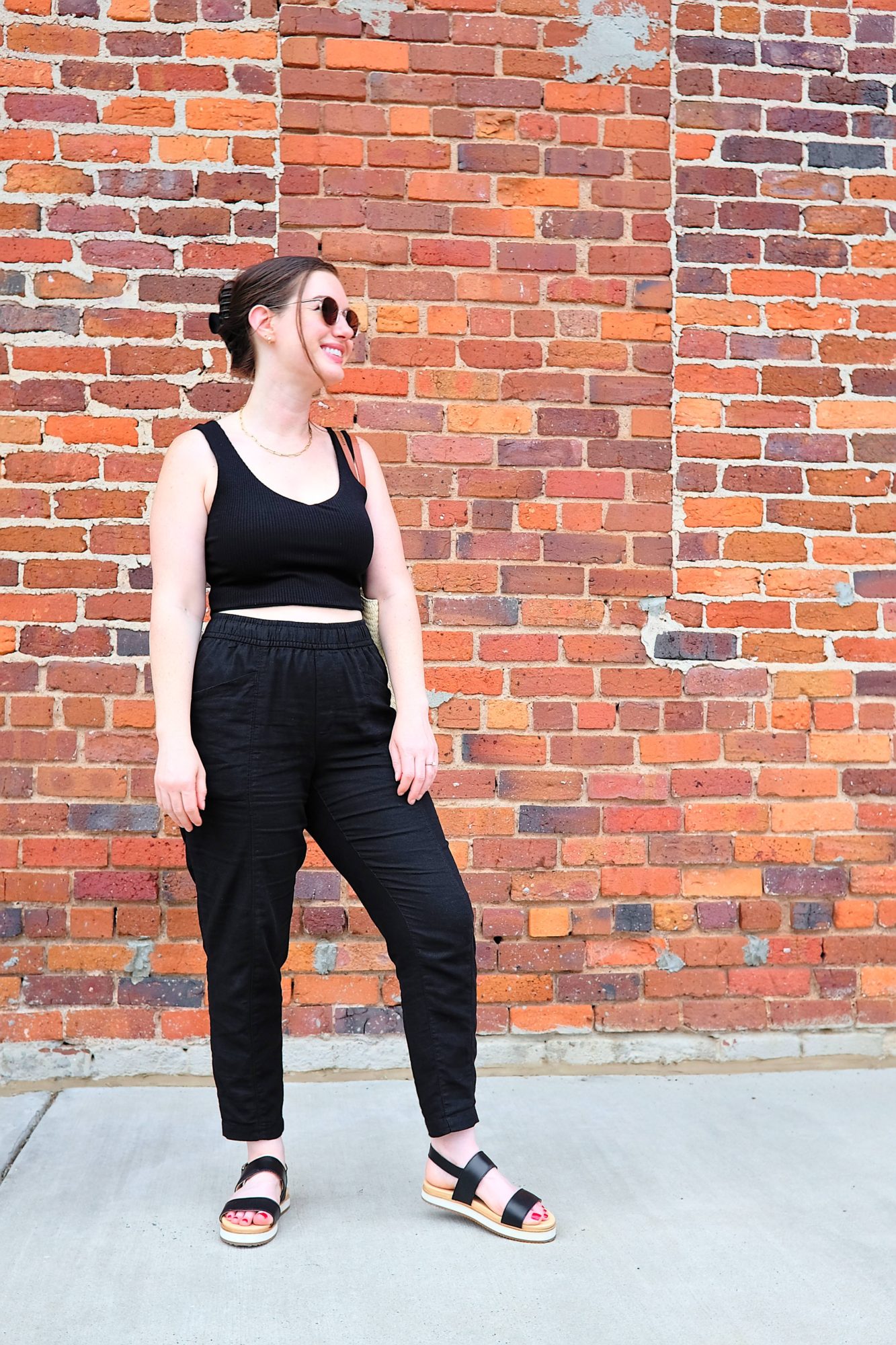 Alyssa stands by a brick wall wearing flatform sandals and linen pants